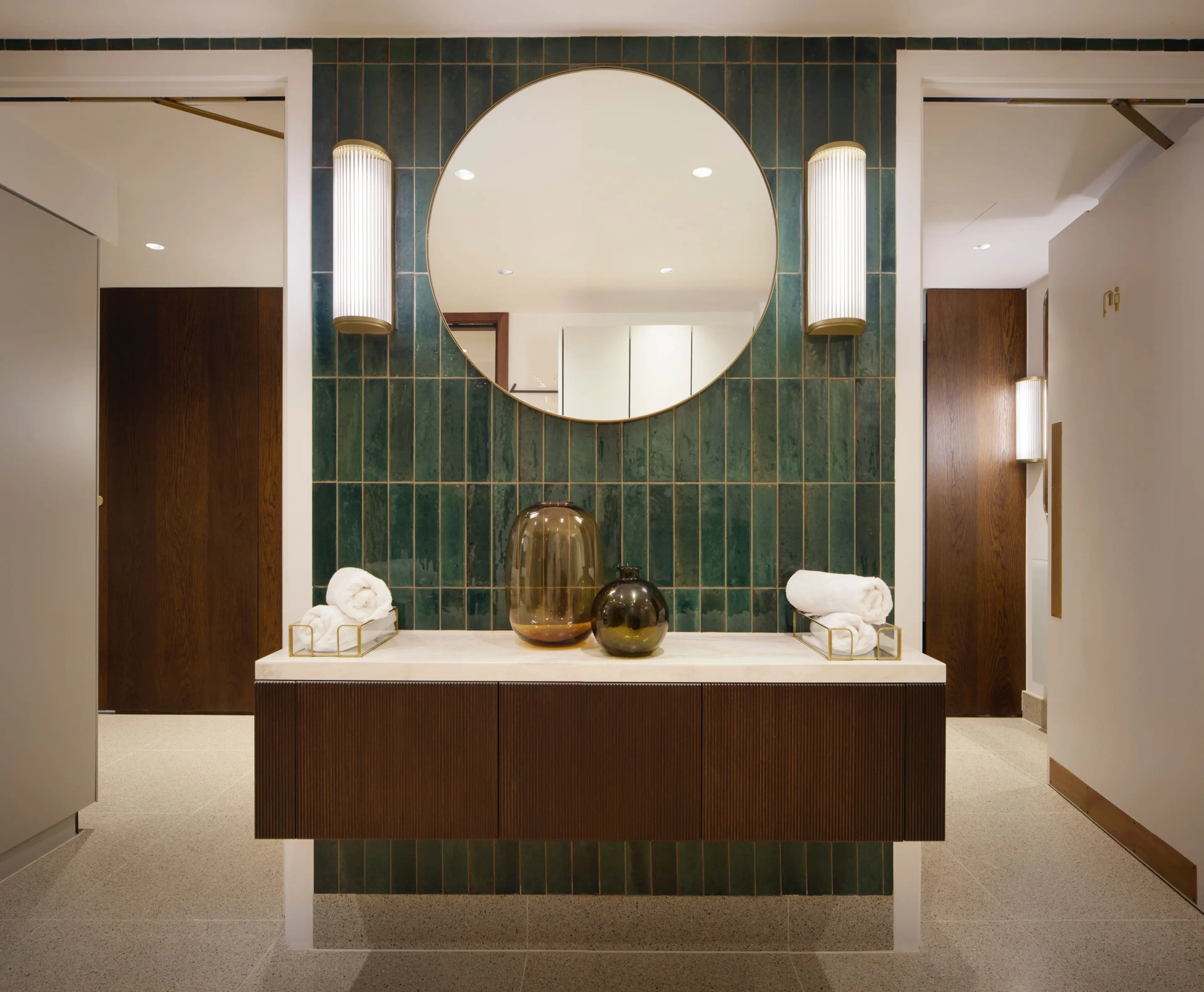 A modern vanity with a round mirror, dark green tiled wall, and dark wood cabinet, flanked by two doorways.