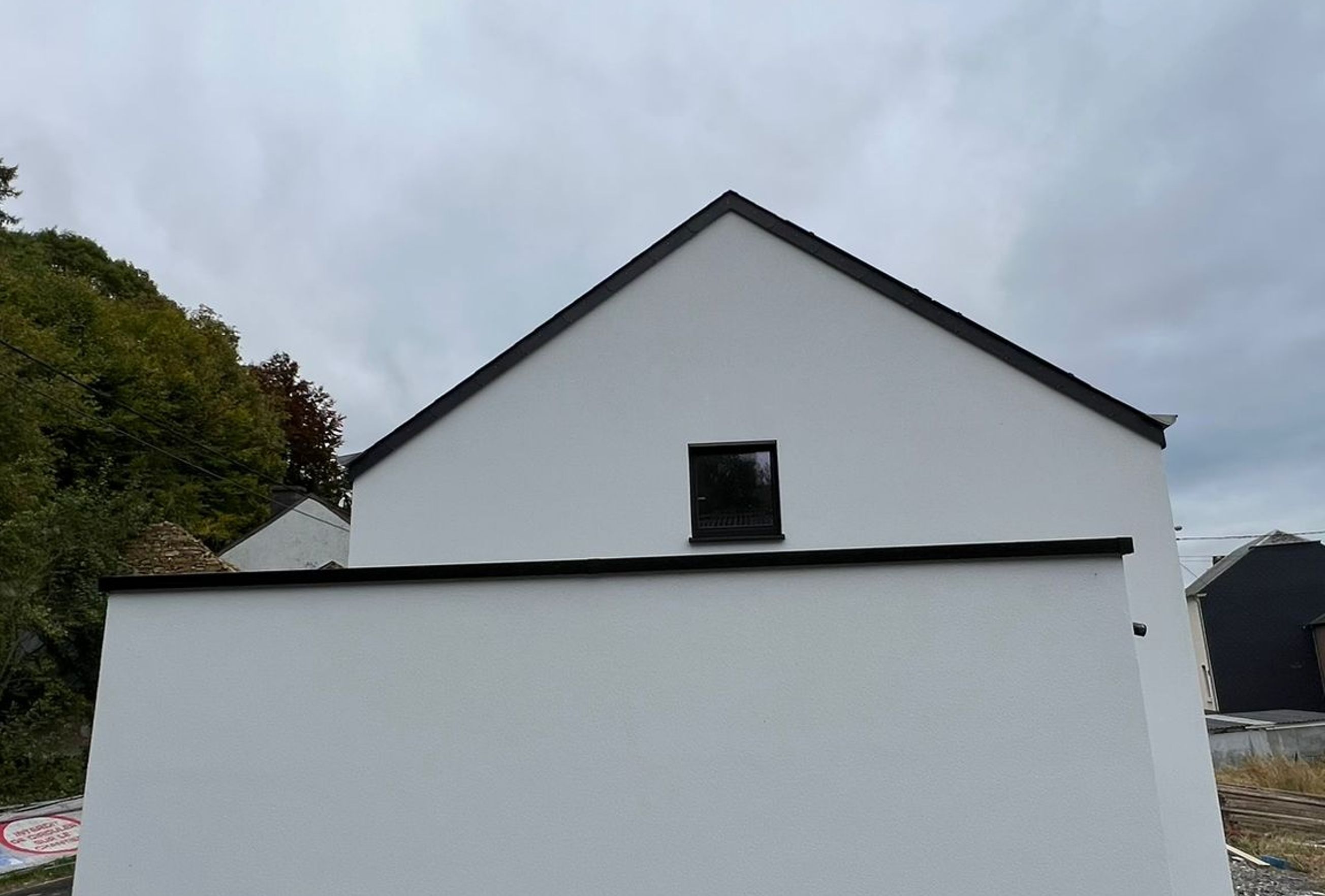 Pignon d’une maison neuve avec façade blanche enduite et petite fenêtre à l’étage, entourée d’un terrain encore en chantier sous un ciel nuageux.