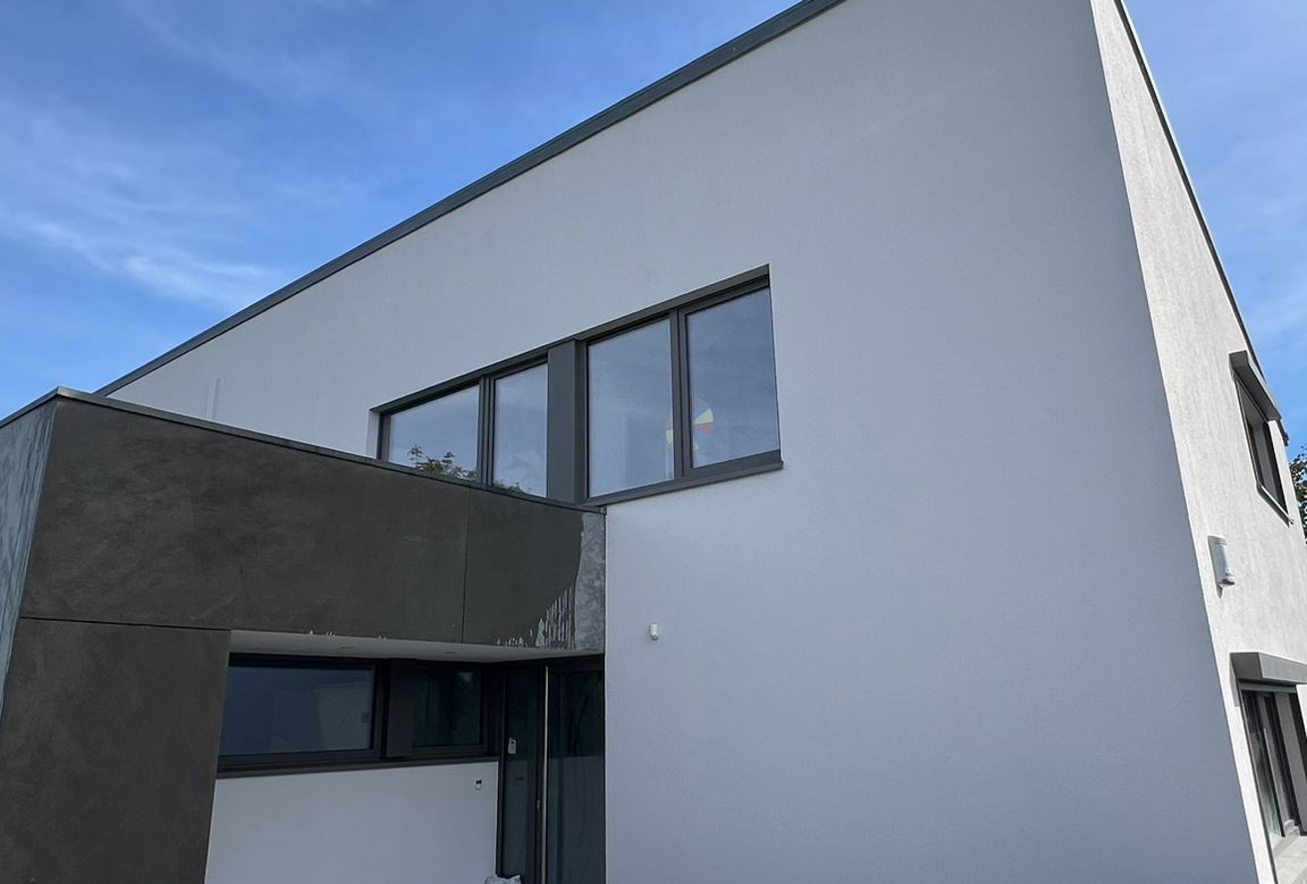 Façade d’une maison contemporaine blanche avec toit incliné et extension sombre, vue sur une terrasse moderne en cours d’aménagement sous un ciel bleu.