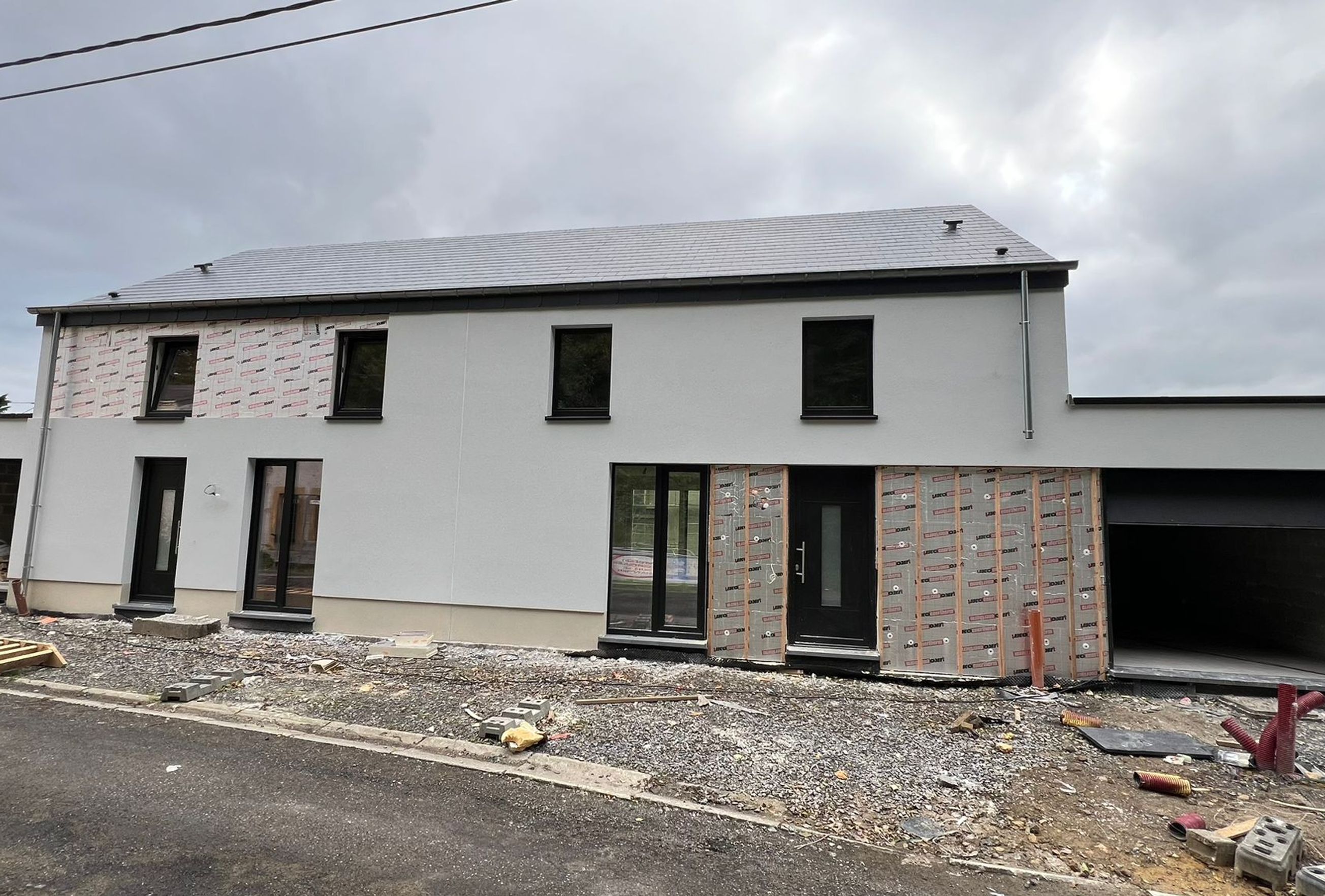 Maison moderne en cours de construction avec façade partiellement enduite, ouvertures pour portes et fenêtres installées et garage intégré, sur un chantier avec gravats et matériaux.