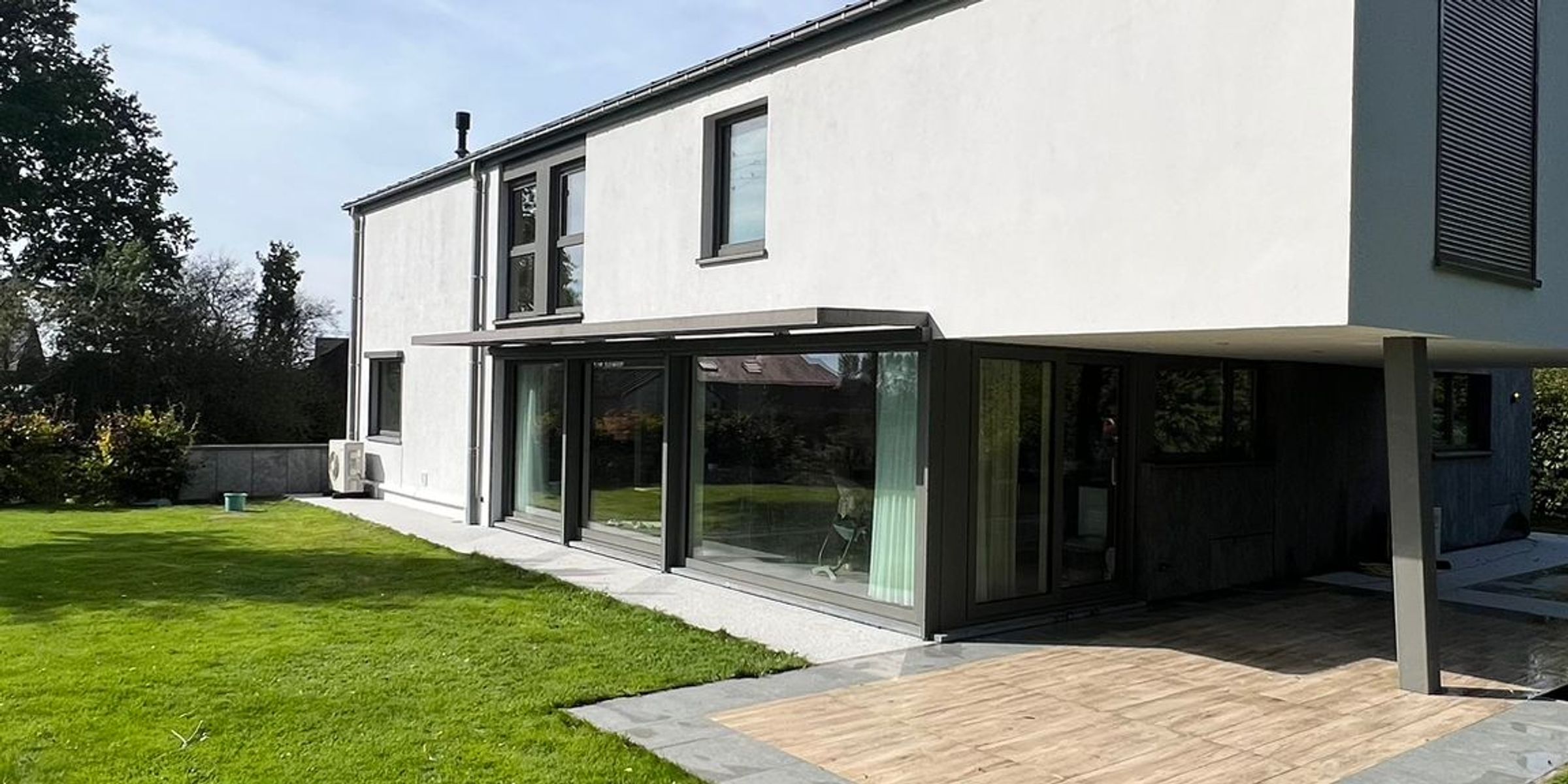Maison moderne avec façade en crépi blanc, grandes baies vitrées et terrasse donnant sur un jardin verdoyant
