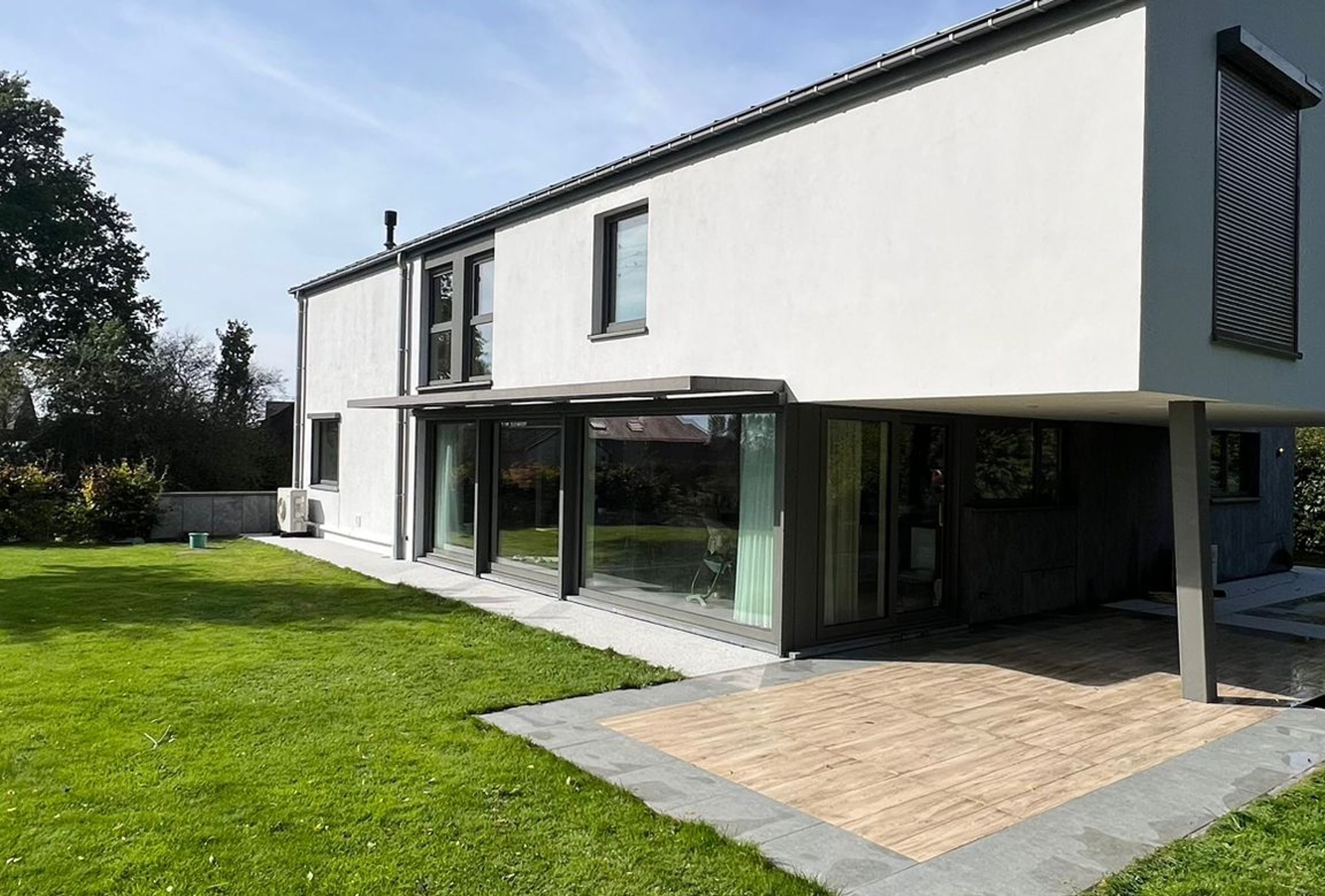 Maison contemporaine blanche à deux étages avec grandes baies vitrées, terrasse couverte et jardin verdoyant sous un ciel dégagé.
