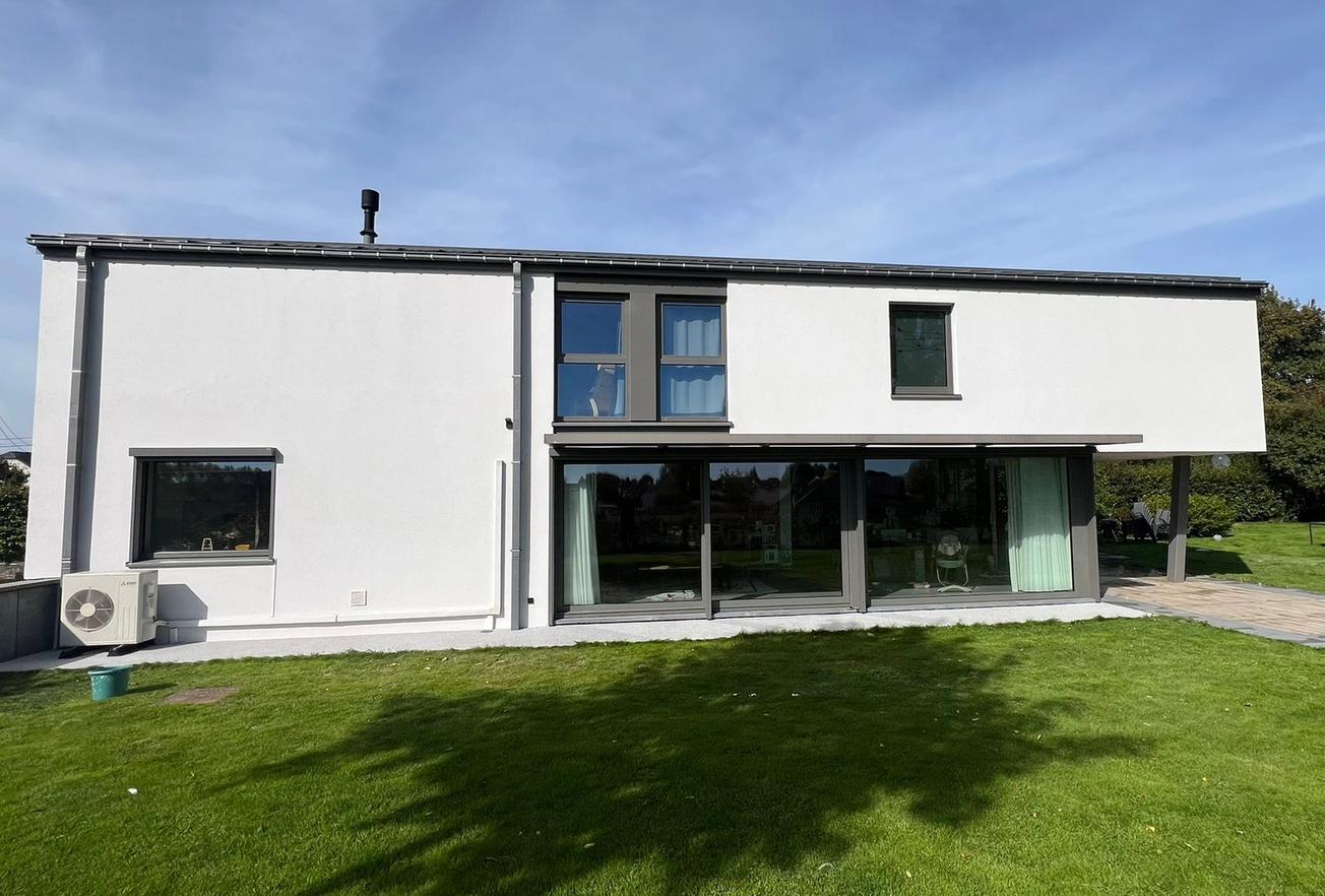 Maison contemporaine à deux étages avec façade blanche, grandes baies vitrées donnant sur le jardin et terrasse couverte, entourée d’une pelouse sous un ciel dégagé.