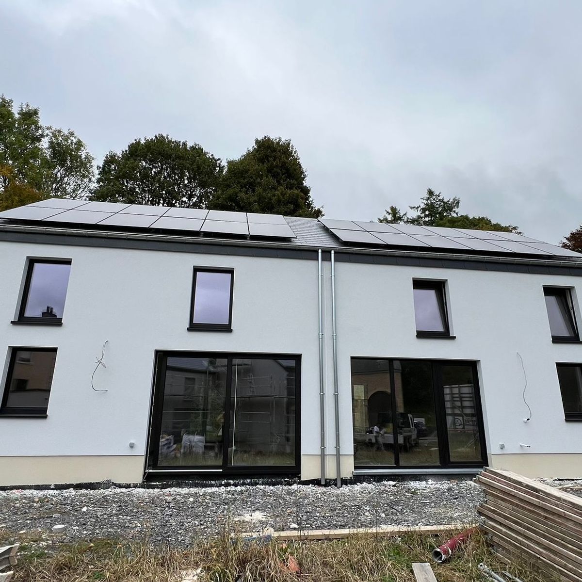 Façade arrière complète d'une maison bi-familiale ou jumelée de style moderne. Elle dispose de larges baies vitrées au rez-de-chaussée, de fenêtres symétriques à l'étage et de panneaux solaires installés sur toute la longueur du toit.