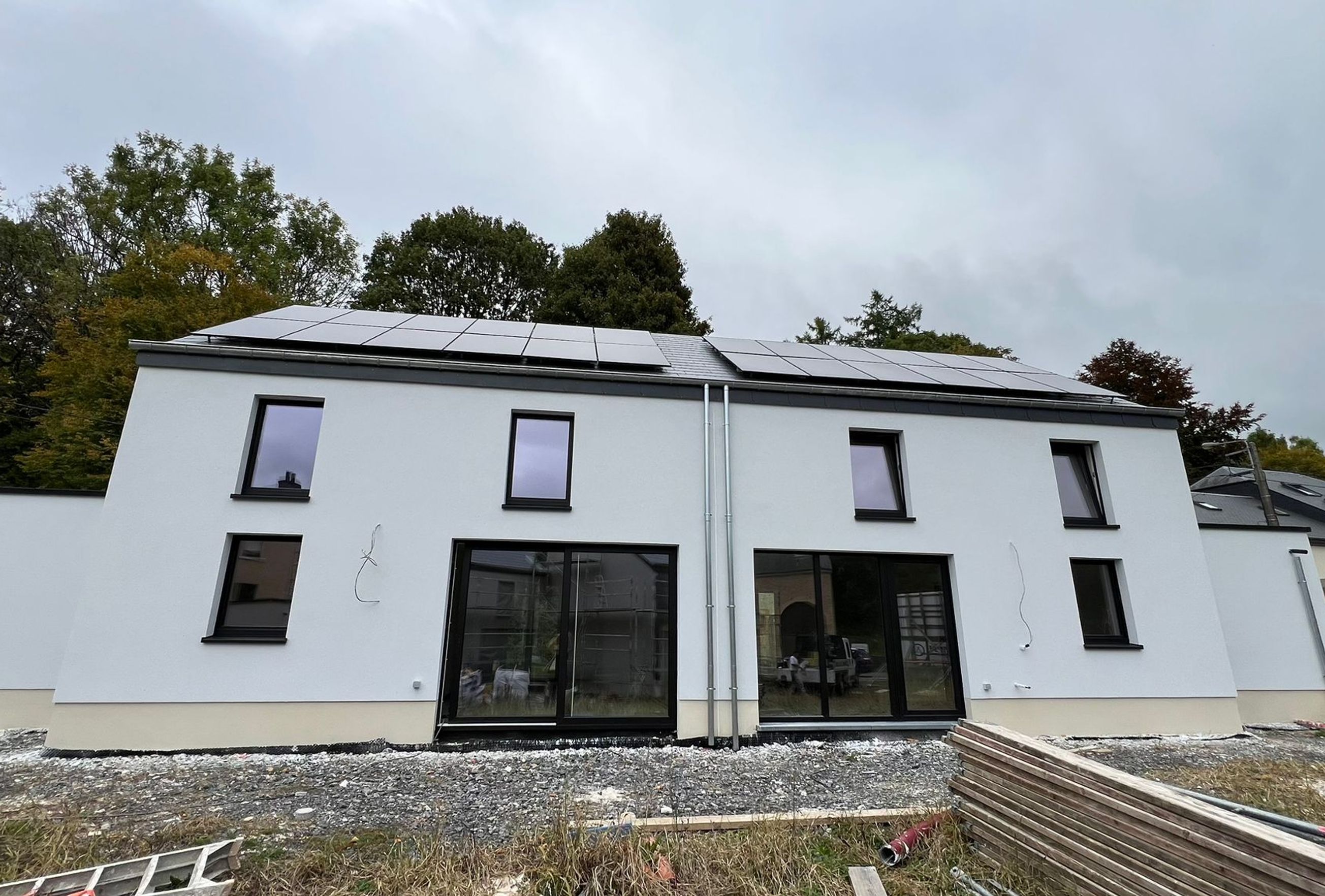 Maison moderne à deux étages avec façade blanche, grandes baies vitrées et panneaux solaires installés sur le toit, sur un terrain encore en chantier.
