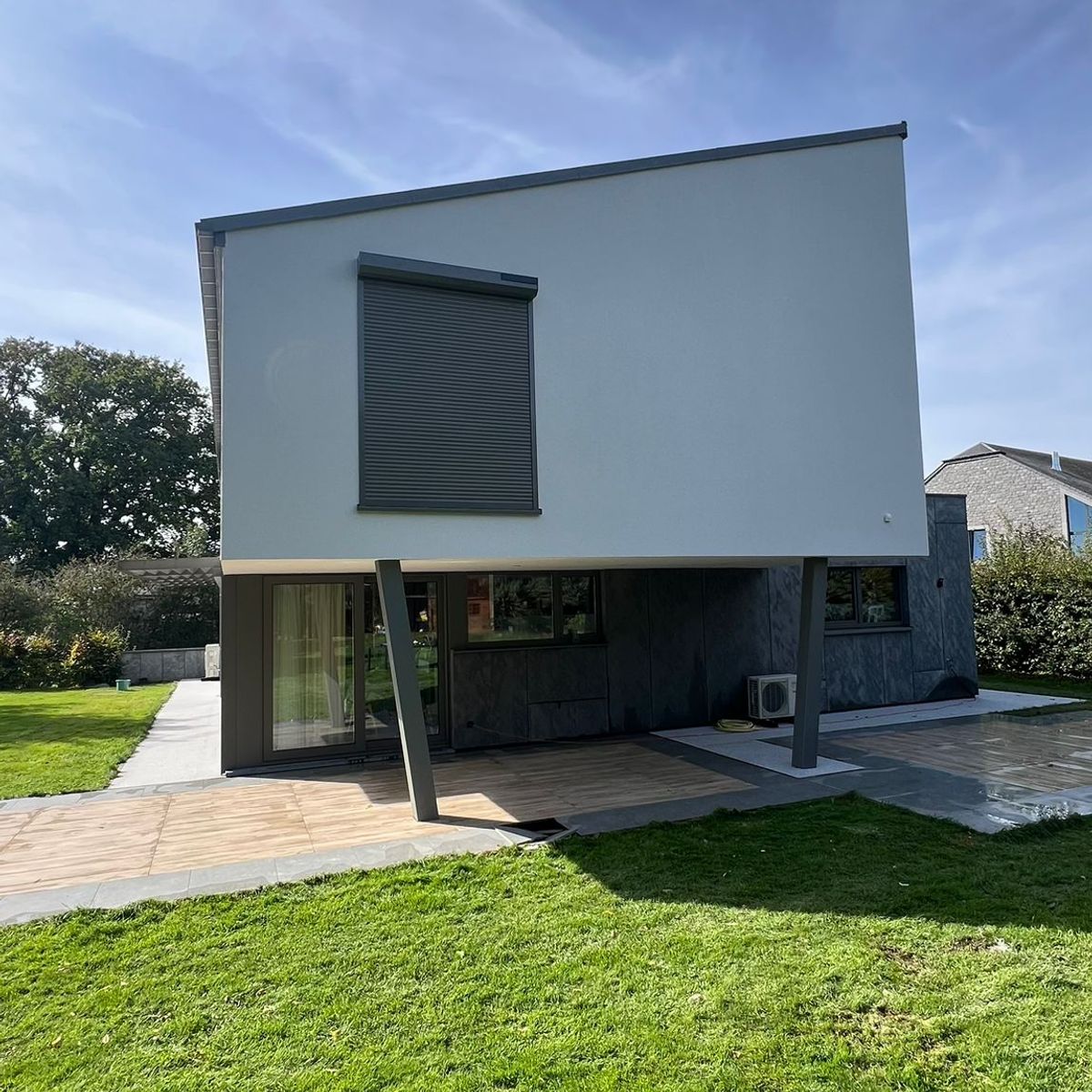 Vue extérieure d'une maison moderne avec un étage supérieur blanc en porte-à-faux soutenu par un pilier incliné. Au rez-de-chaussée, une terrasse en bois jouxte une pelouse verdoyante sous un ciel bleu dégagé.