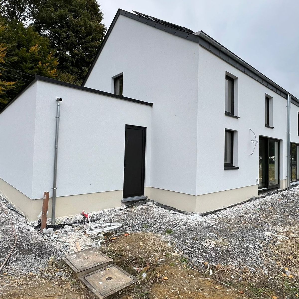 Vue d'une maison bicolore (blanc et soubassement beige) montrant une extension à toit plat et des menuiseries extérieures noires. Le terrain est encore en cours d'aménagement.