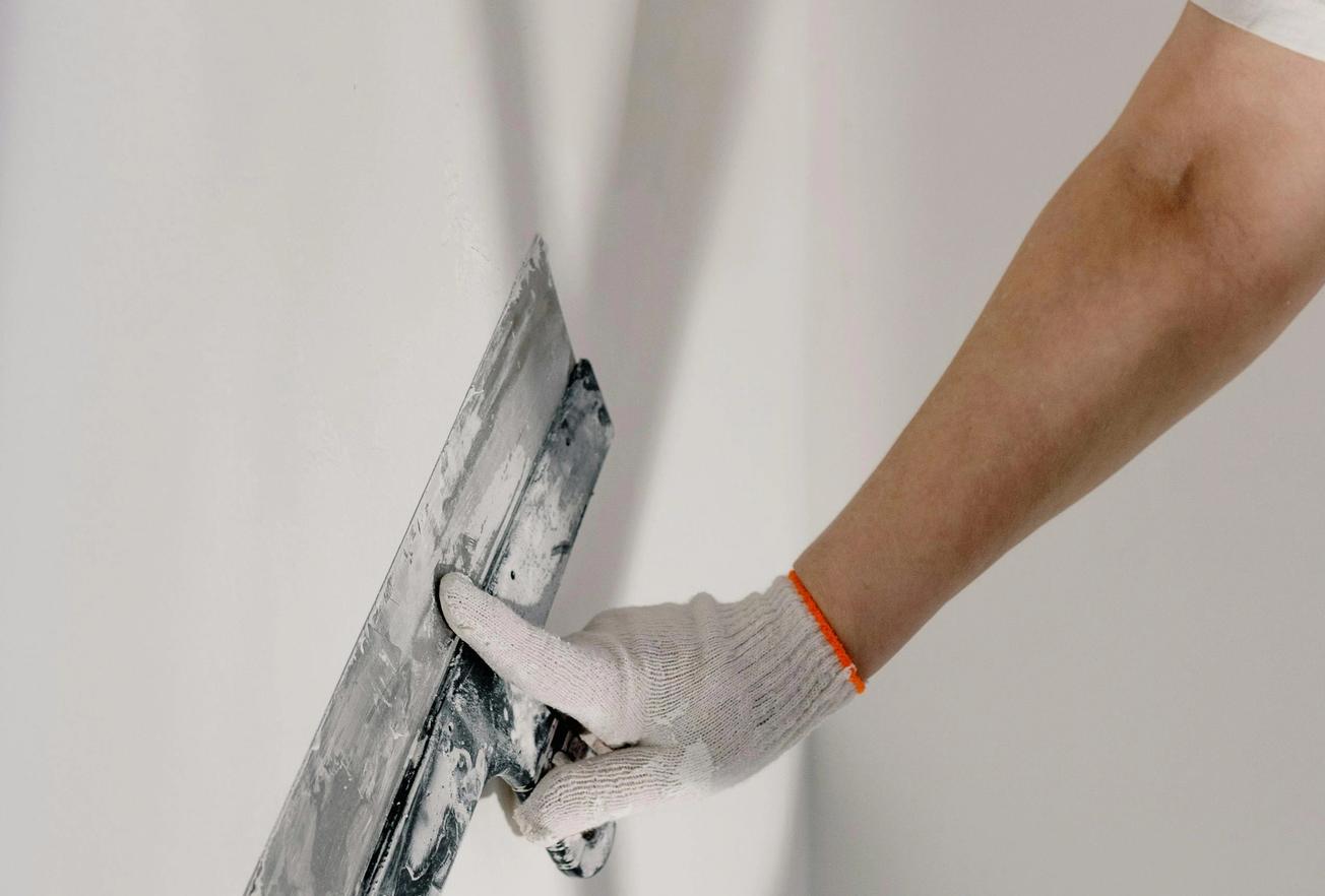 Main gantée appliquant de l’enduit sur un mur blanc à l’aide d’une spatule, lors de travaux de finition intérieure.