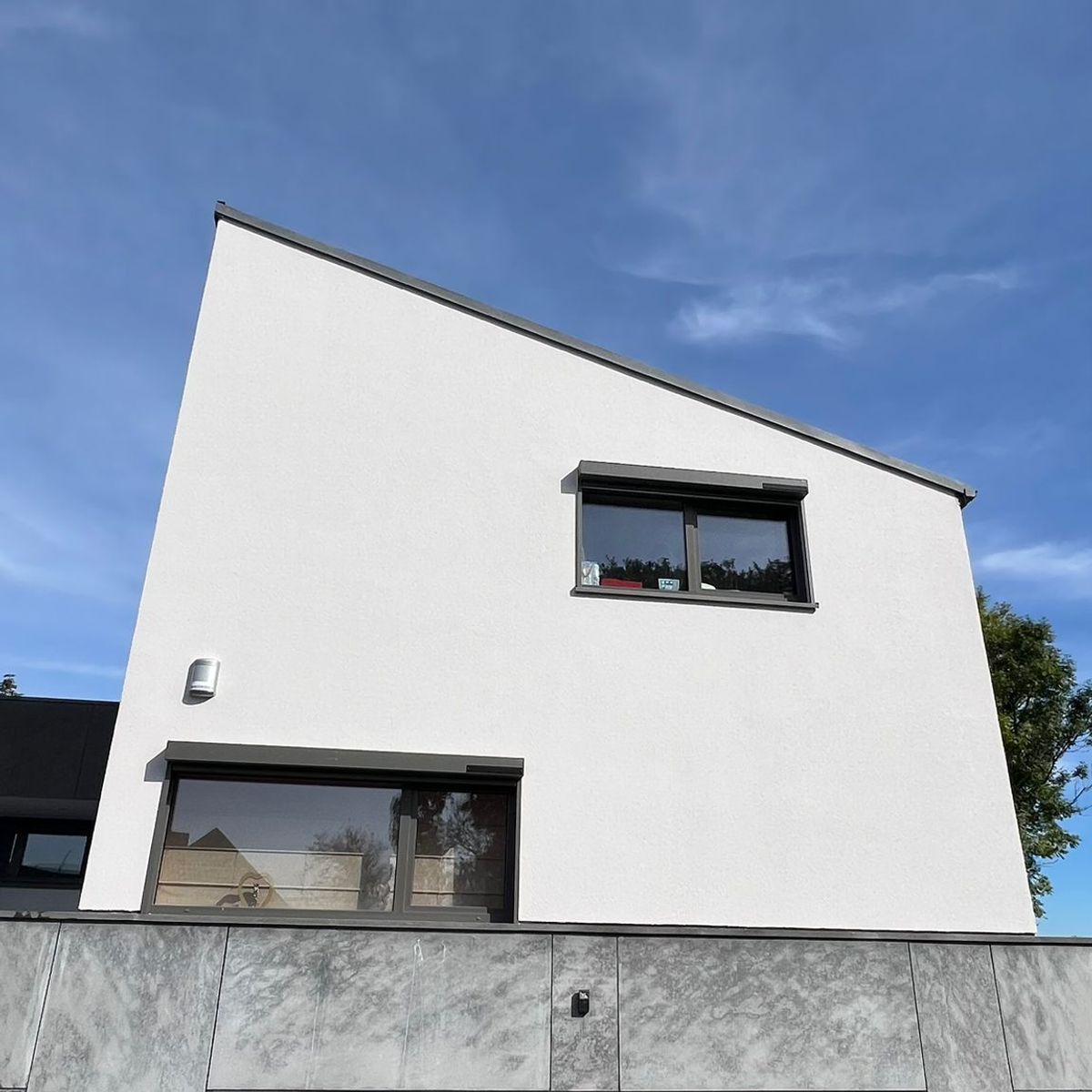 Gros plan sur une section de maison moderne présentant un rez-de-chaussée avec un revêtement en pierre grise texturée et une porte de garage anthracite. L'étage supérieur est en crépi blanc avec un toit en pente monopente.