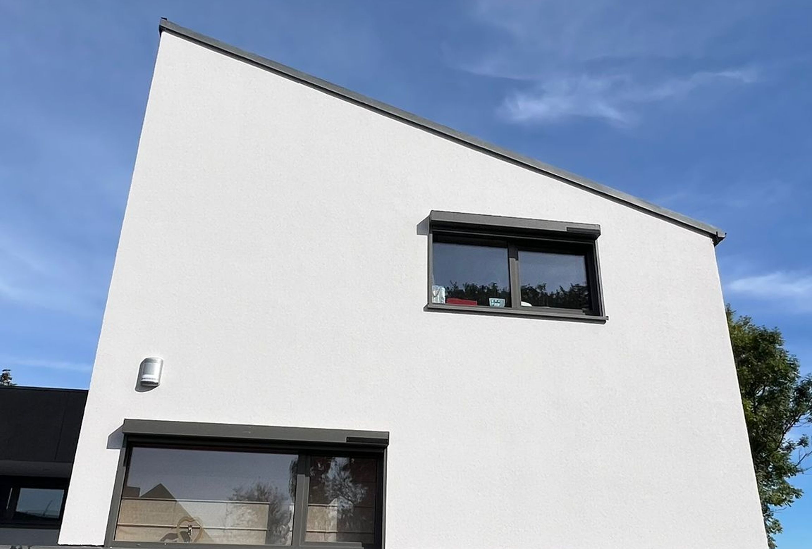 Façade d’une maison contemporaine avec étage enduit blanc au-dessus d’un garage intégré habillé de panneaux gris, sous un ciel bleu dégagé.