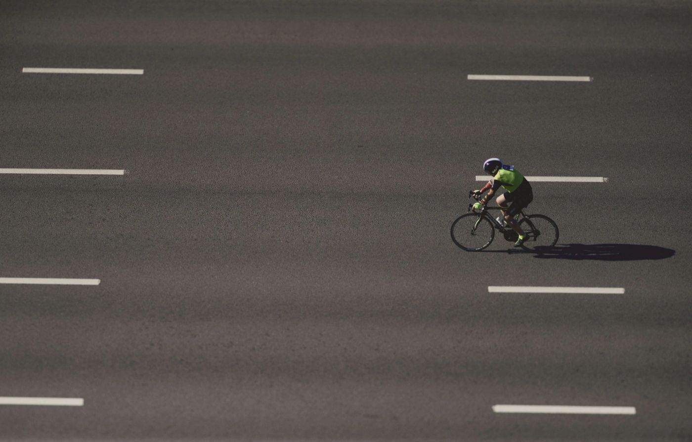 Aerial view of a cyclist in bright green athletic wear riding across a dark asphalt surface marked with white lane dividers, casting a long shadow while moving through the geometric pattern of road markings, symbolizing choice, determination, and purposeful movement forward