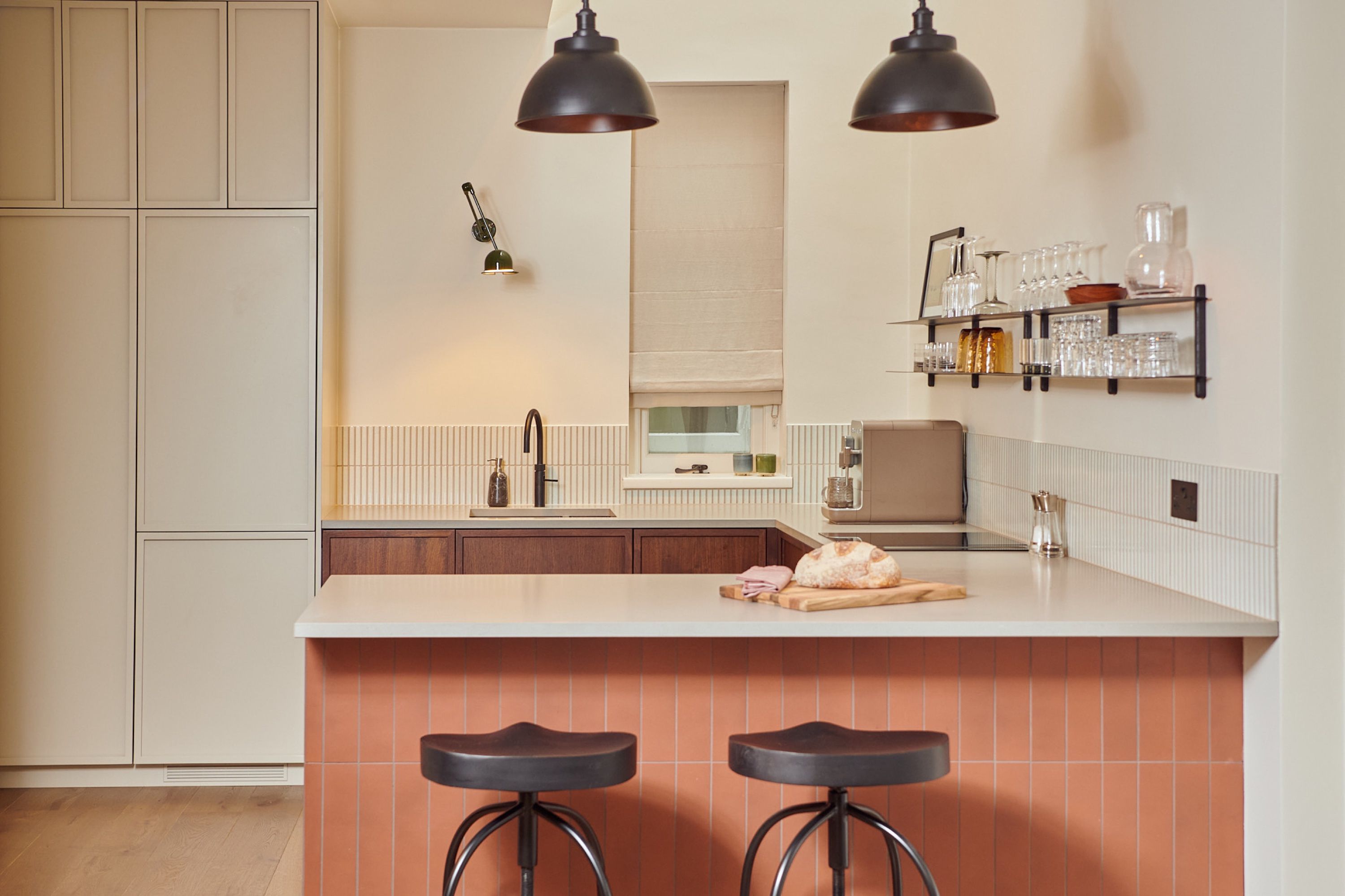 Kitchen with two black tractor stools at the terracotta tiled peninsula, walnut units behind and an articulated brass wall light on the left wall.