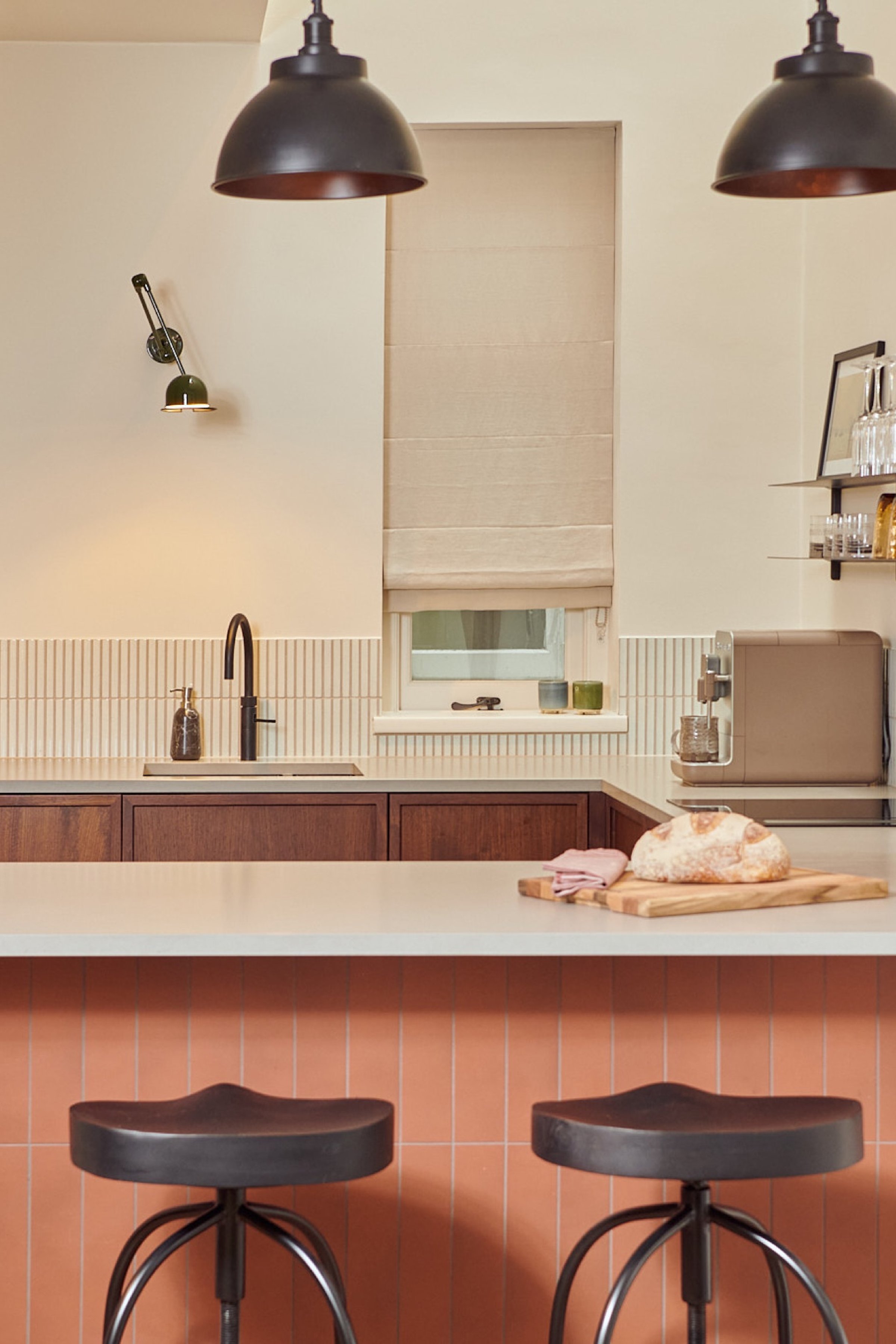 Kitchen with two black tractor stools at the terracotta tiled peninsula, walnut units behind and an articulated brass wall light on the left wall.