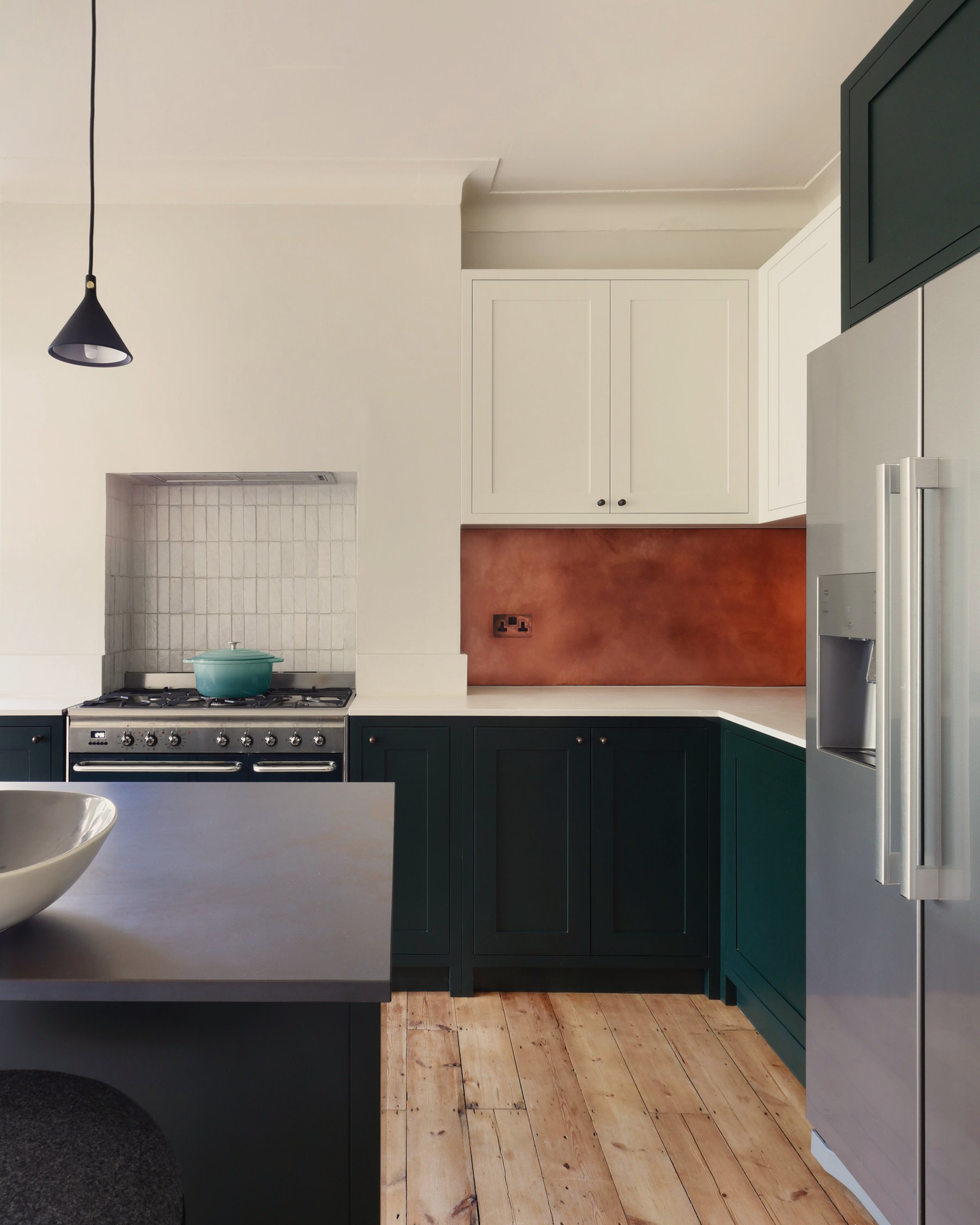 Kitchen corner in dark green and off-white, with a range cooker in a tiled recess, a patinated copper splashback and cream wall cabinets above.