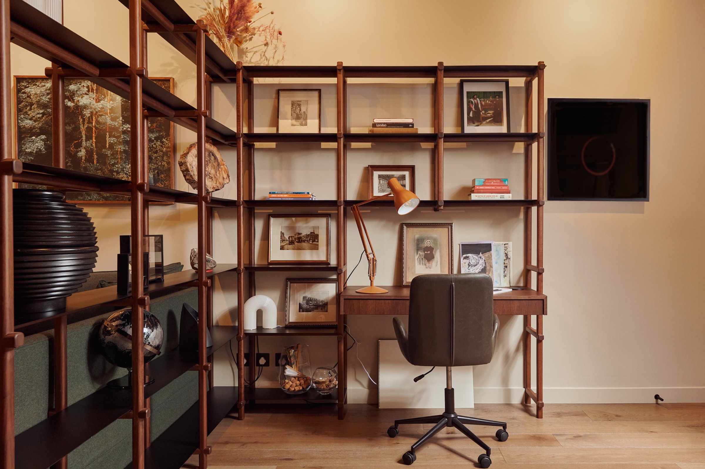 Study shelving seen wider, its timber uprights carrying framed pictures, books and objects around the integrated desk and orange lamp.