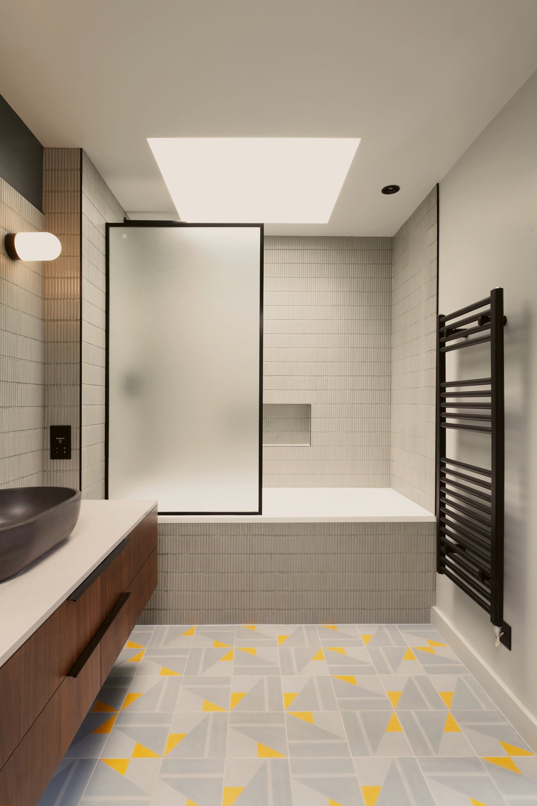 The bath end of the bathroom under a rooflight, with a black-framed fluted glass screen, fluted white tiling and a patterned grey and yellow floor.