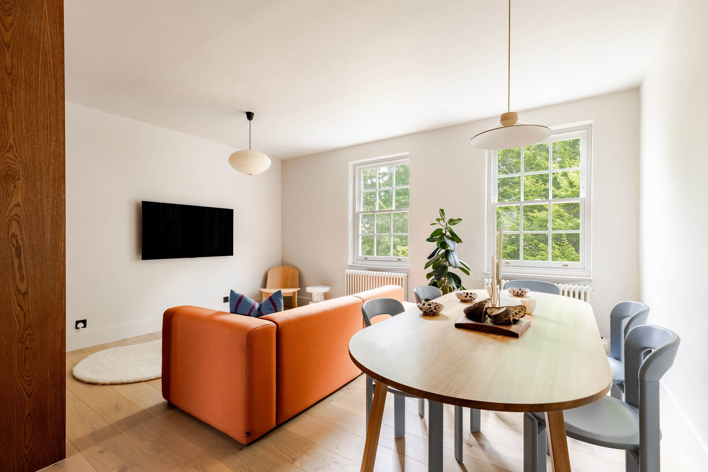 Living and dining room with an orange sofa, an oval dining table under a pendant, and two sash windows onto trees.