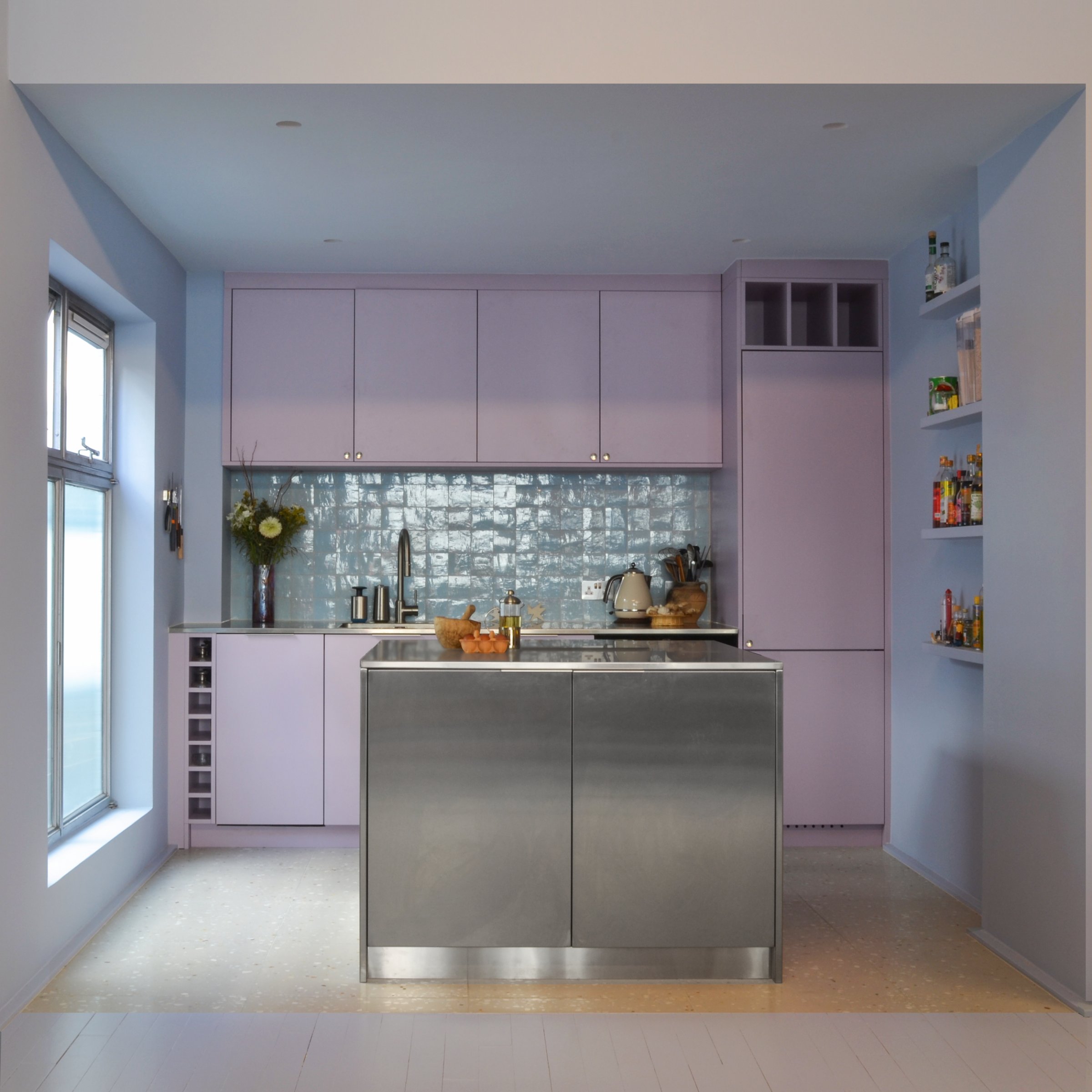 Lilac kitchen with a glazed blue-green tile splashback, a stainless steel island and a wine rack built into the end of the run