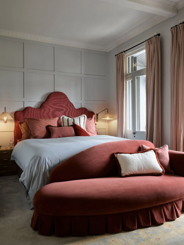 Bedroom with a scalloped burgundy headboard, burgundy cushions and a curved burgundy daybed at the foot of the bed, against pale panelled walls