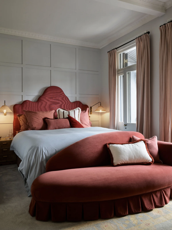 Bedroom with a scalloped burgundy headboard, burgundy cushions and a curved burgundy daybed at the foot of the bed, against pale panelled walls