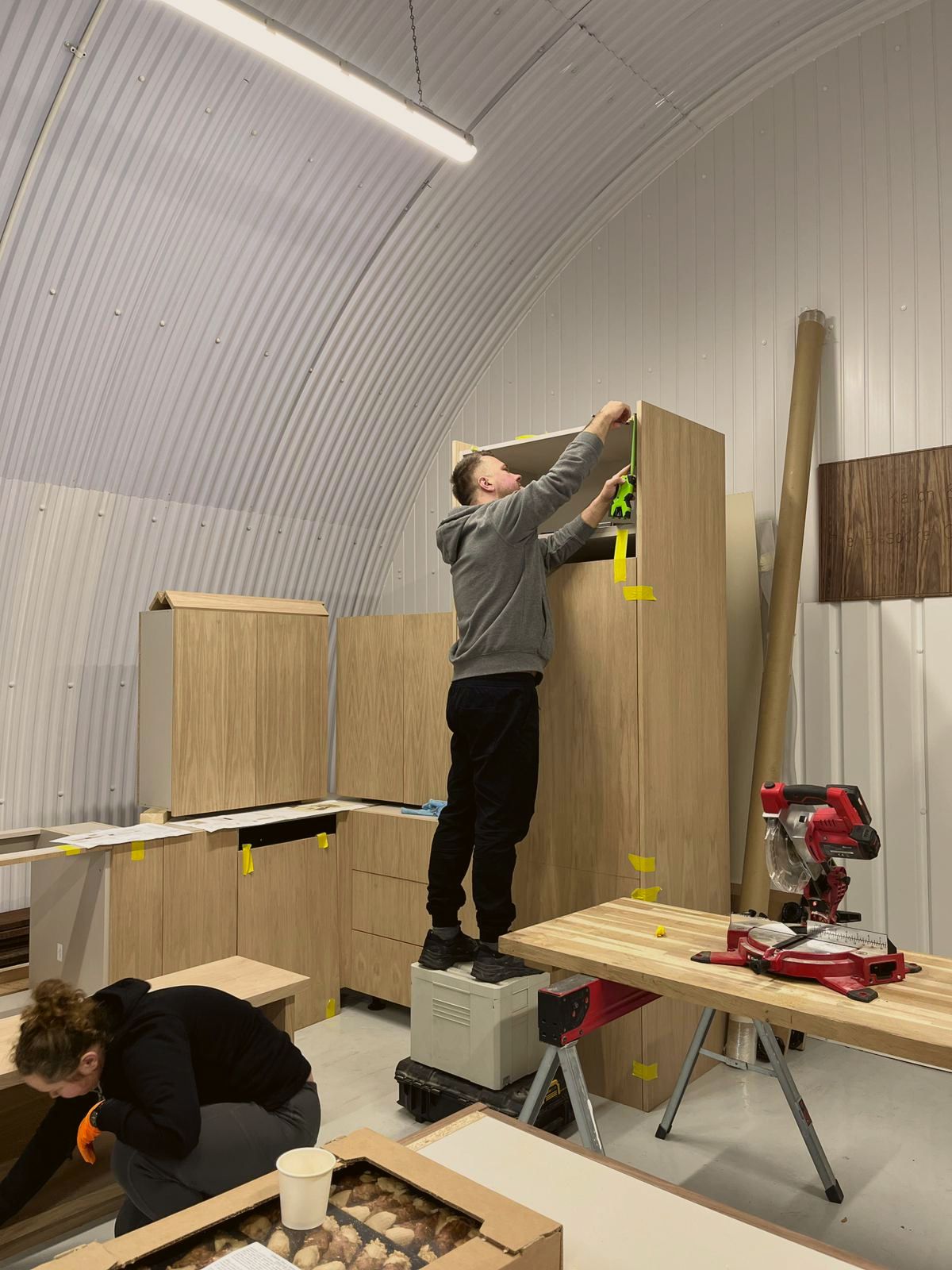 In the arched workshop, a joiner on a step fixes the top of a tall oak cabinet while another works on a base unit, a mitre saw on the bench beside them