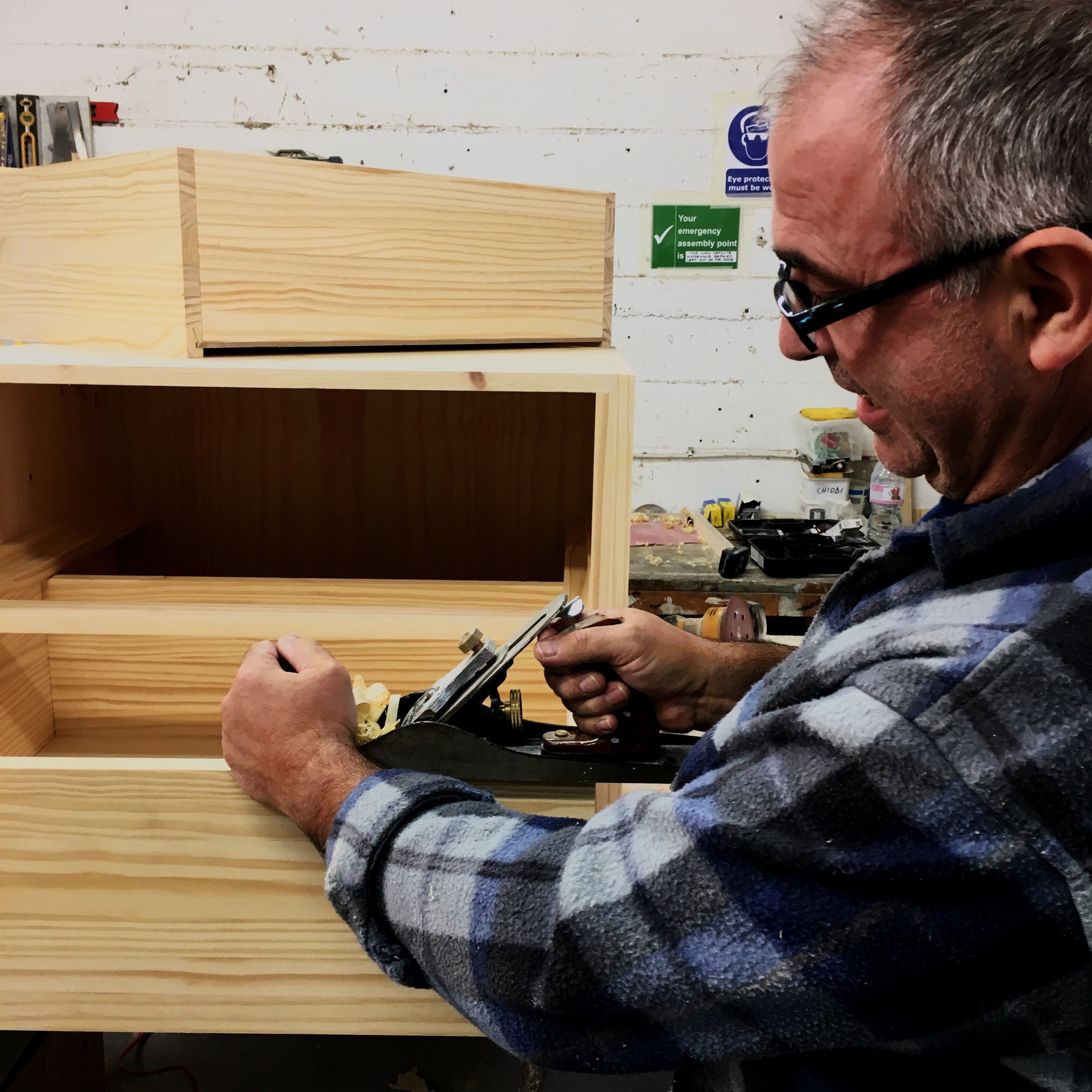 A joiner in safety glasses planing the edge of a pine carcass at the workshop bench.