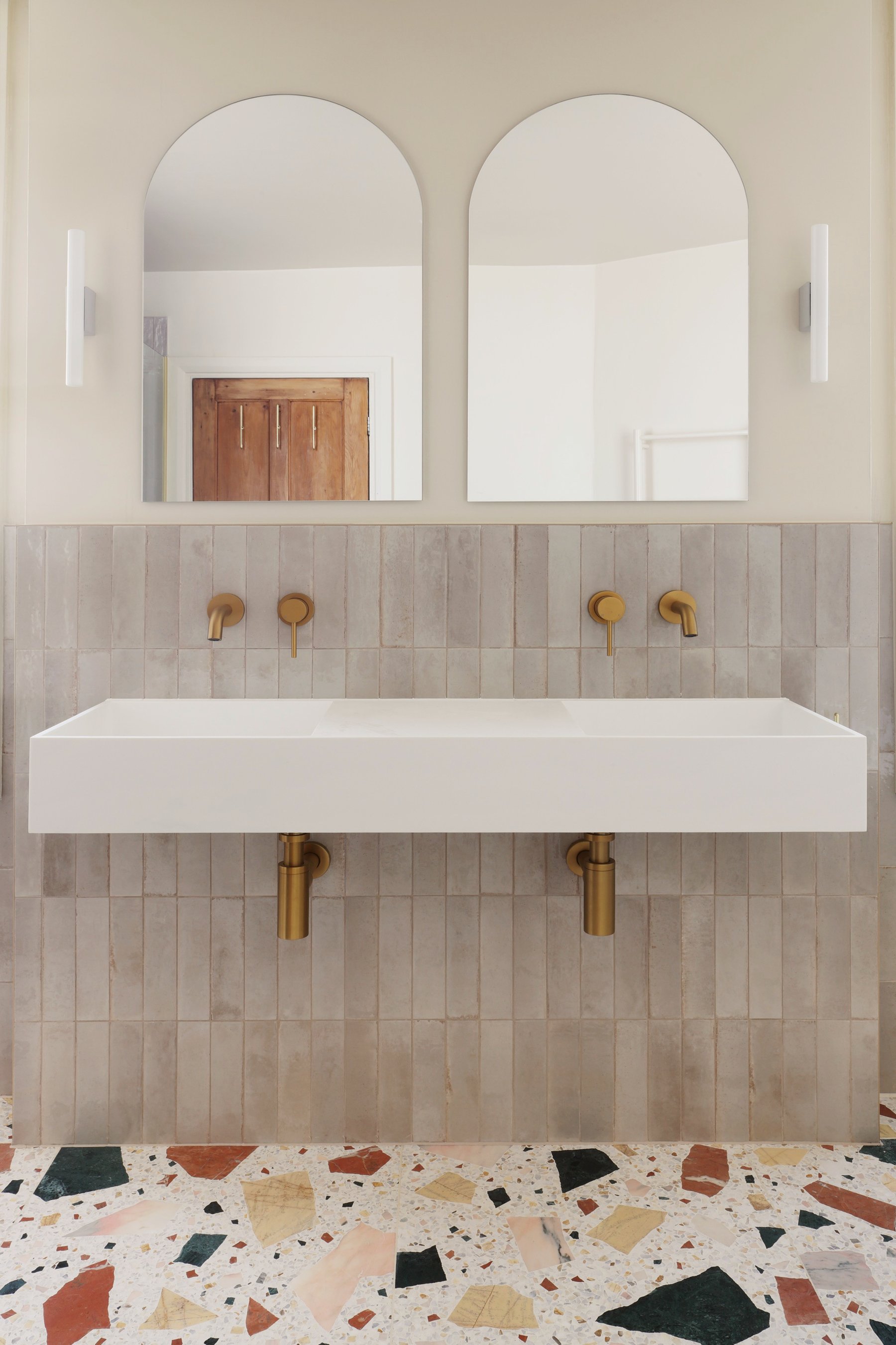 Bathroom vanity: a long white trough basin wall-hung over vertical pale tiles, with brass mixers and exposed traps below two arched frameless mirrors.