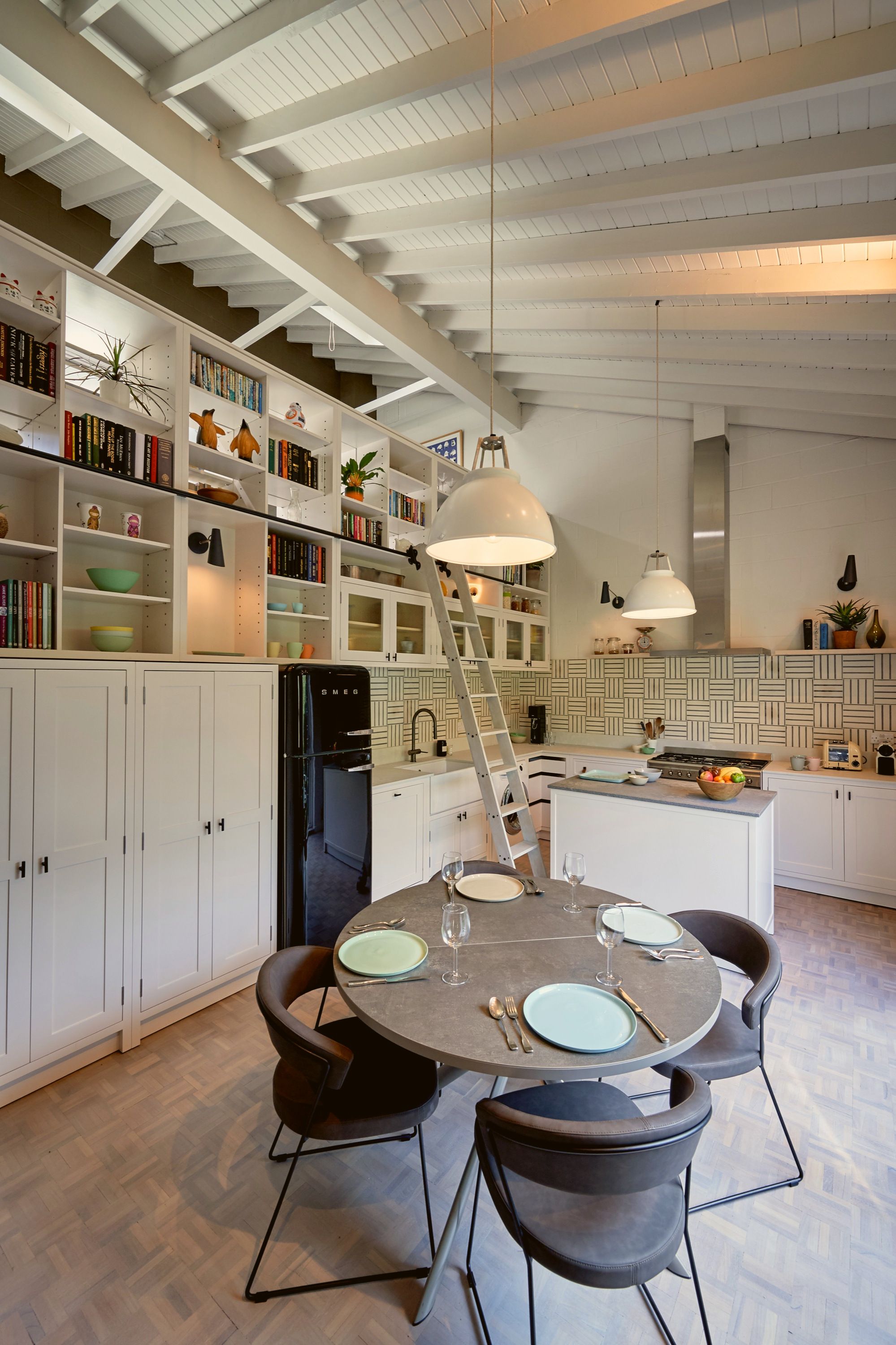 Kitchen and dining space under a white boarded pitched roof, joinery and library ladder left, range and island centre, round table laid for four.