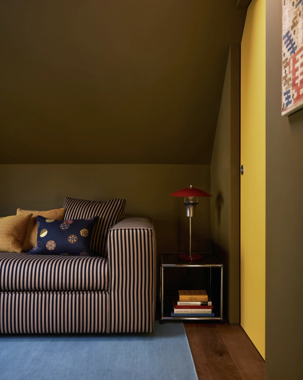 Attic room painted entirely in olive green, with a navy and cream striped sofa, a red table lamp and a yellow door