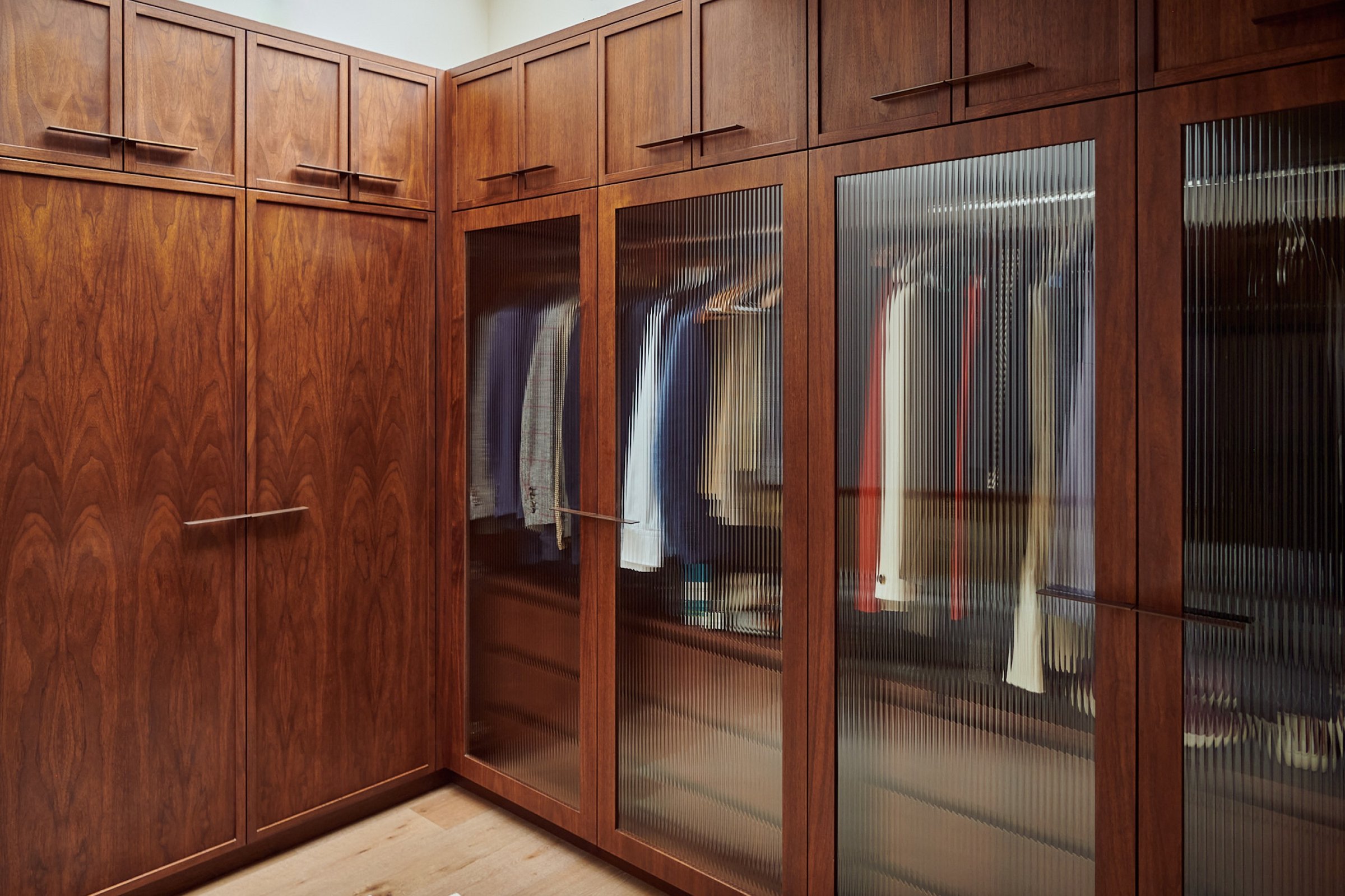 Dressing room corner where solid walnut wardrobe doors turn into reeded glass fronts, the clothes behind softened to colour and shadow.
