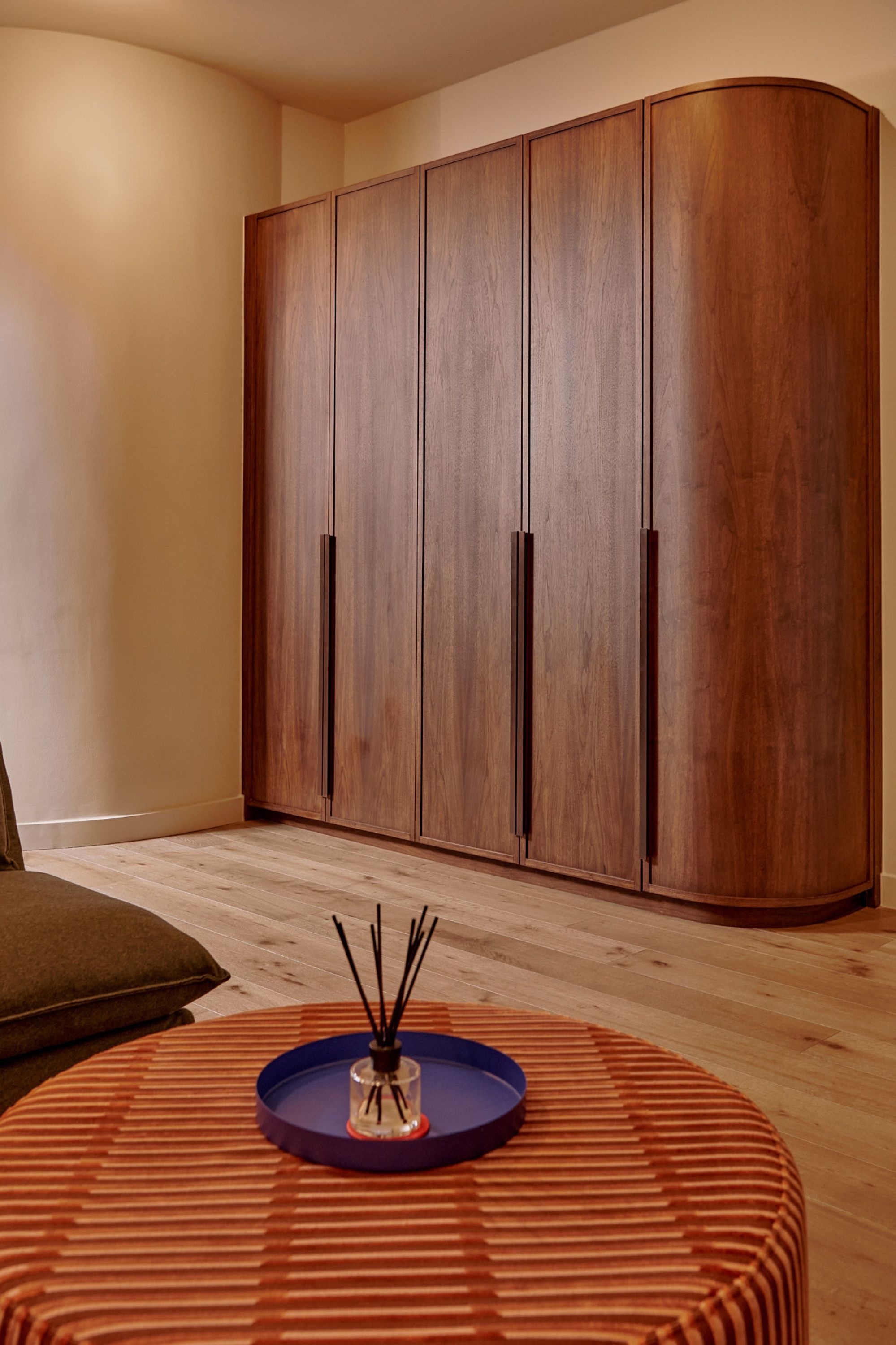 Bedroom wardrobes in walnut with slim dark handles, the end unit curved to meet a rounded plaster wall, past a ribbed rust ottoman.