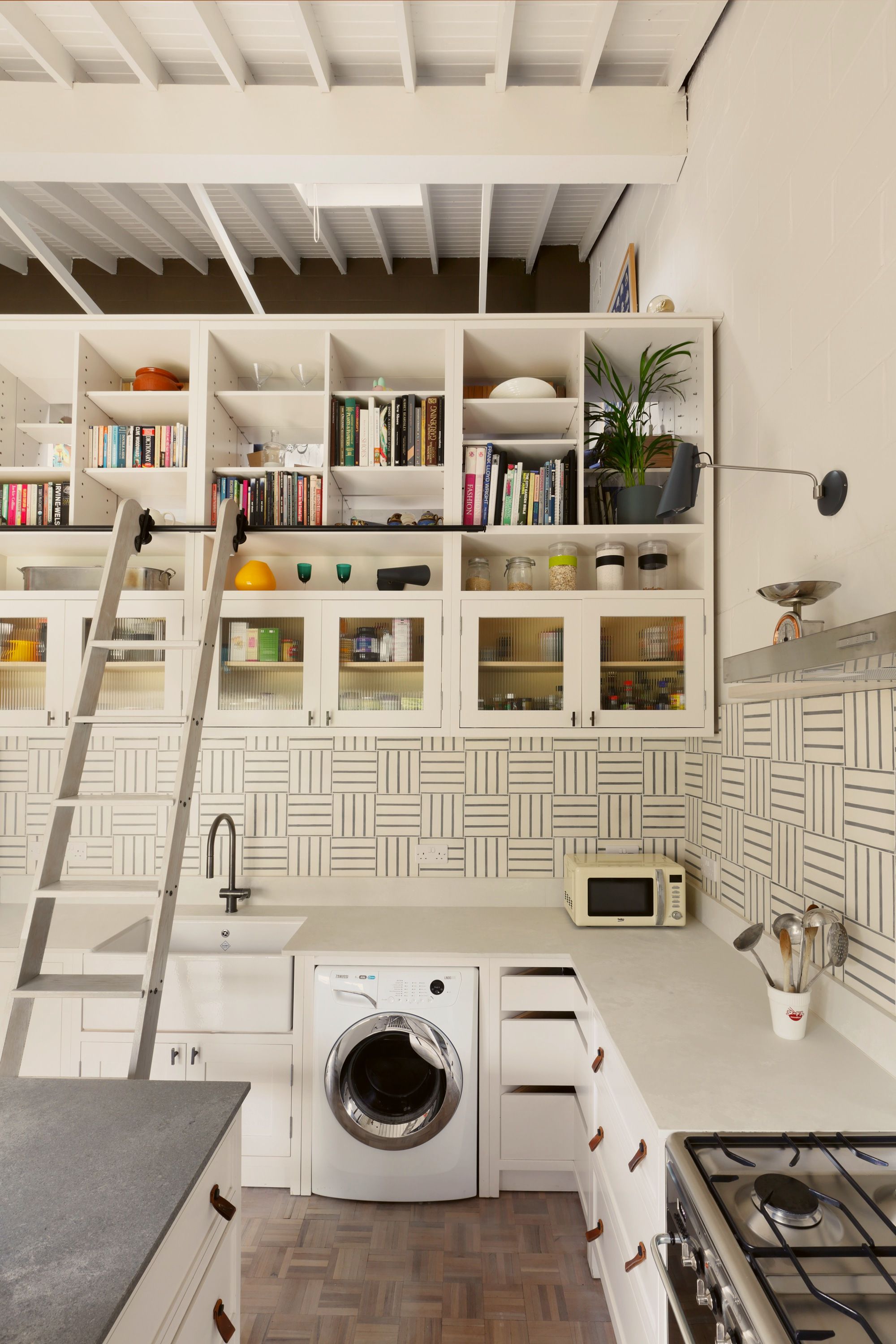 Wall of white joinery rising into a boarded pitched ceiling, with glazed cupboards, open shelves of books and a library ladder on a rail.