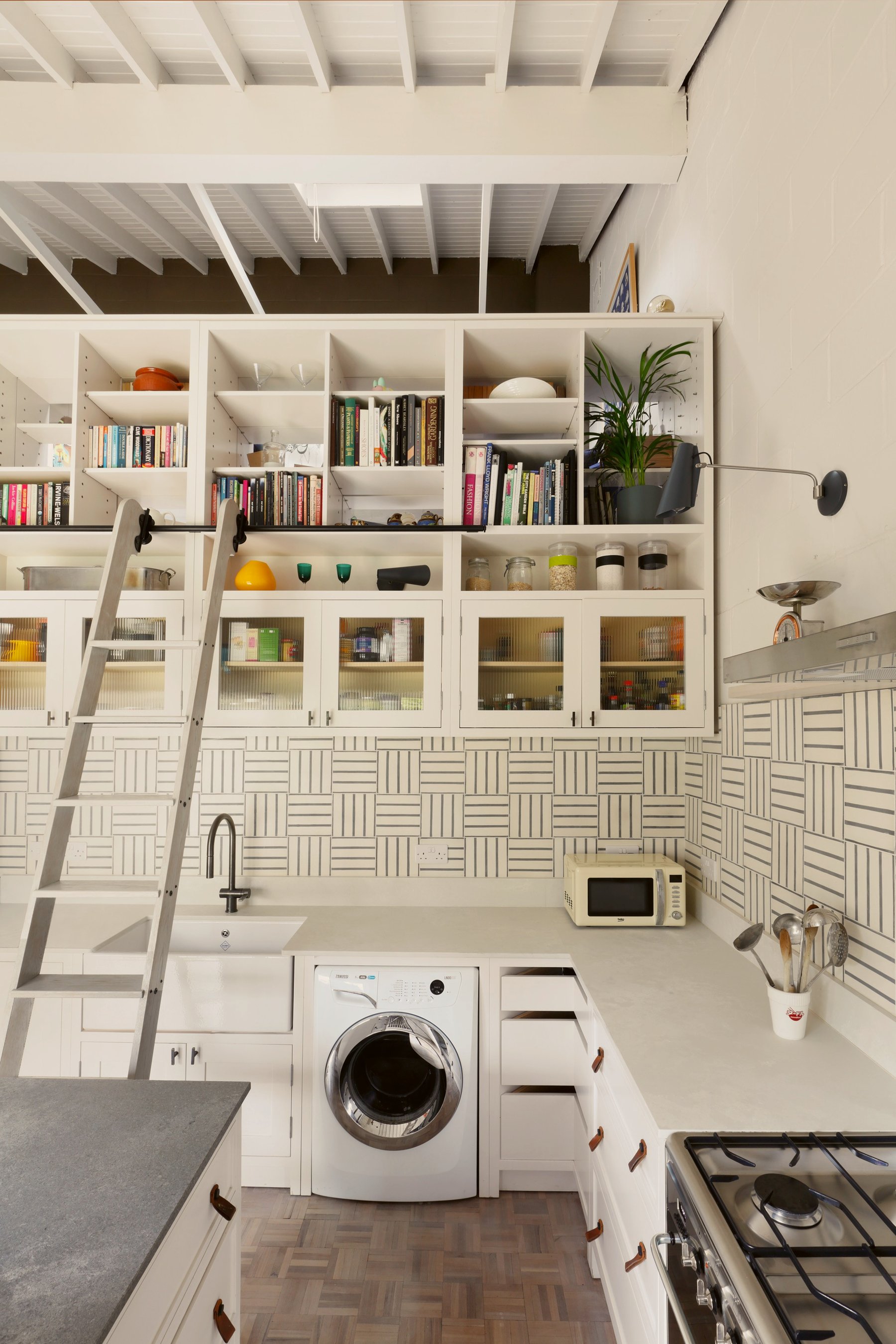 Wall of white joinery rising into a boarded pitched ceiling, with glazed cupboards, open shelves of books and a library ladder on a rail.
