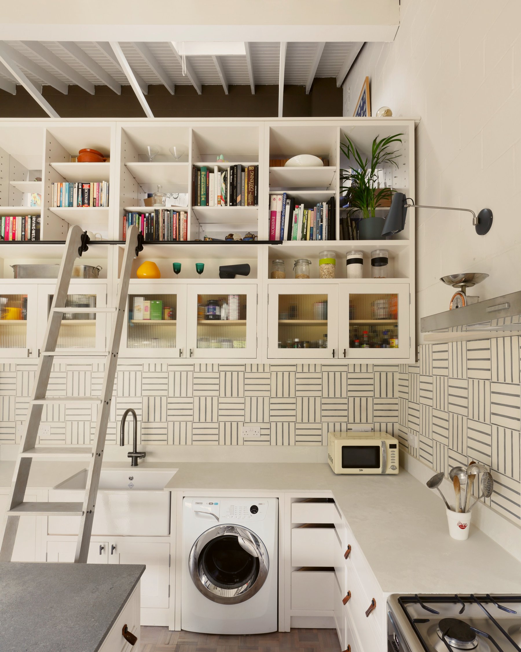 Wall of white joinery rising into a boarded pitched ceiling, with glazed cupboards, open shelves of books and a library ladder on a rail.