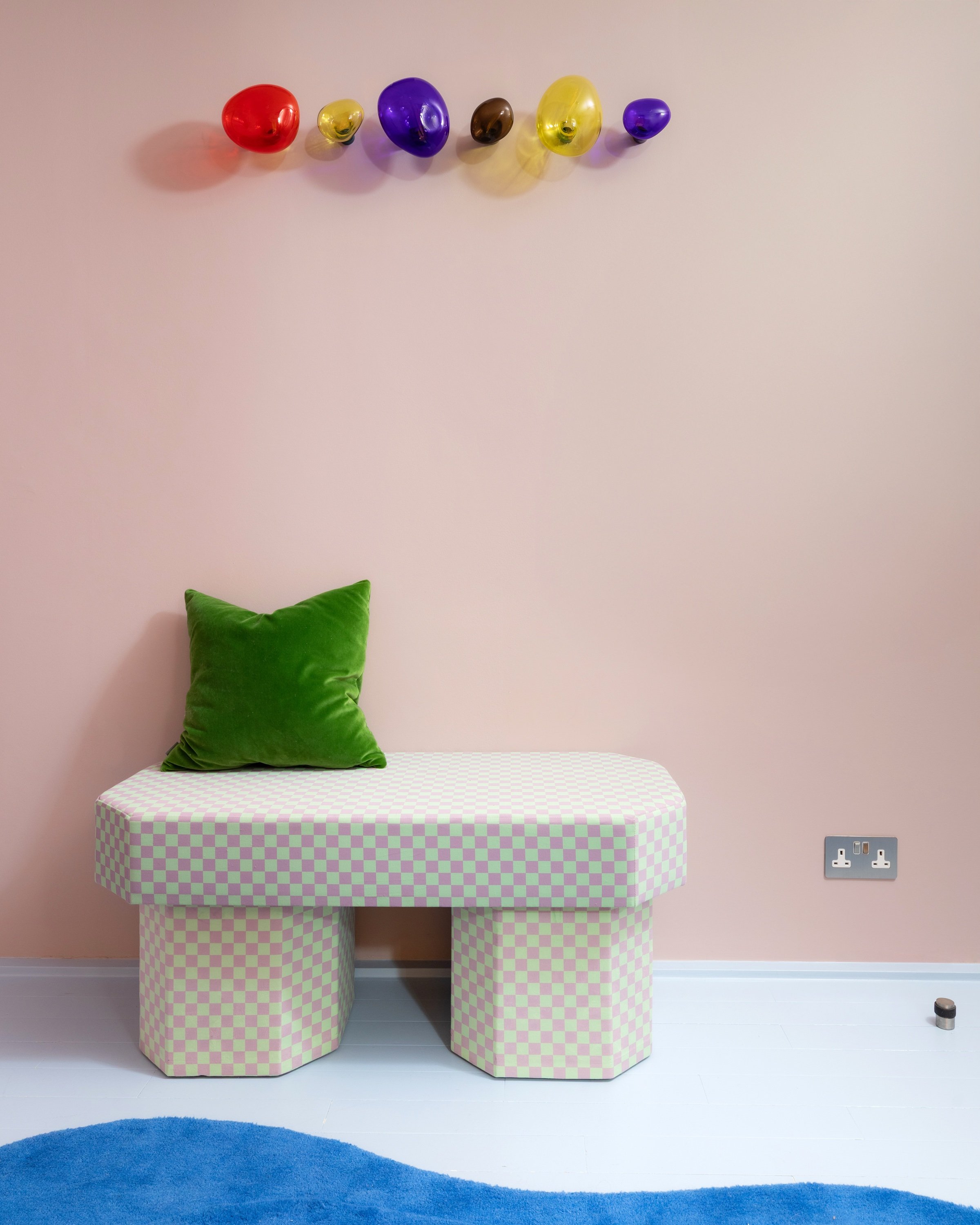 Hallway bench upholstered in pale green and pink checkerboard, with a green velvet cushion and a row of coloured glass wall hooks on a soft pink wall.