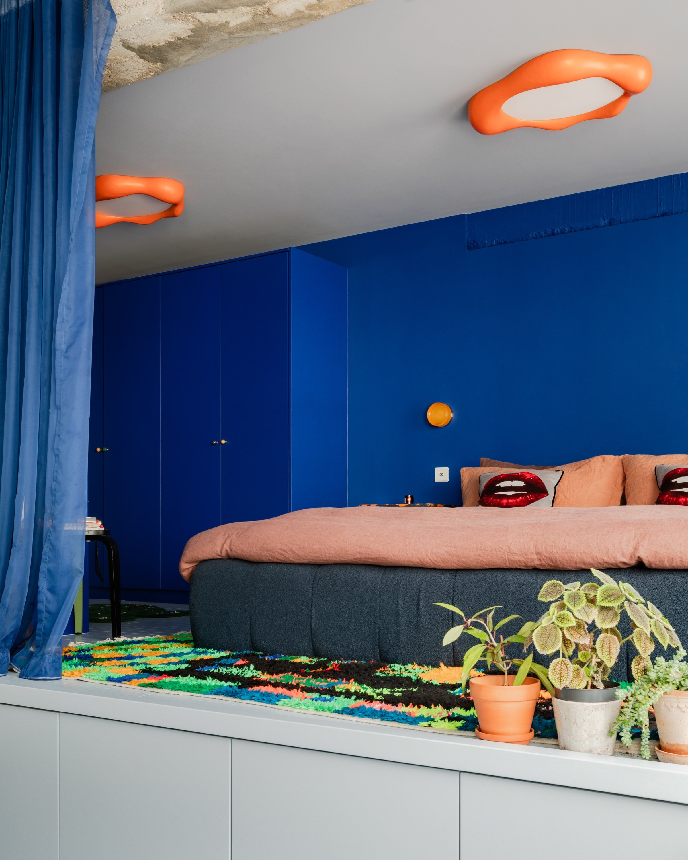 The sleeping platform from the side, blue velvet curtain drawn back, orange ceiling lights against rough concrete, deep blue fitted wardrobes behind.