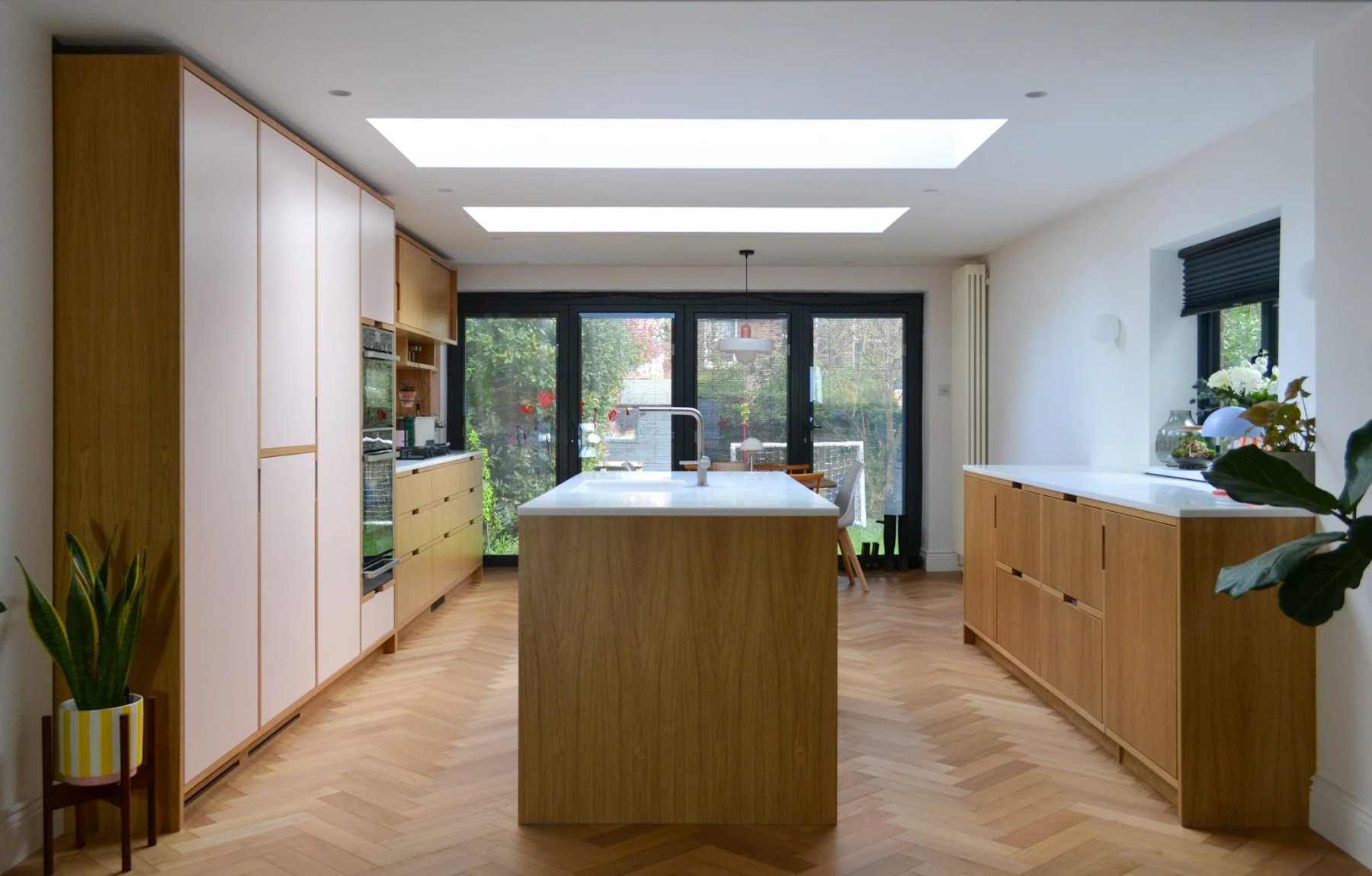 Open-plan kitchen and dining room with an oak island, full-height pale pink cabinets, herringbone parquet and skylights over bifold doors to the garden