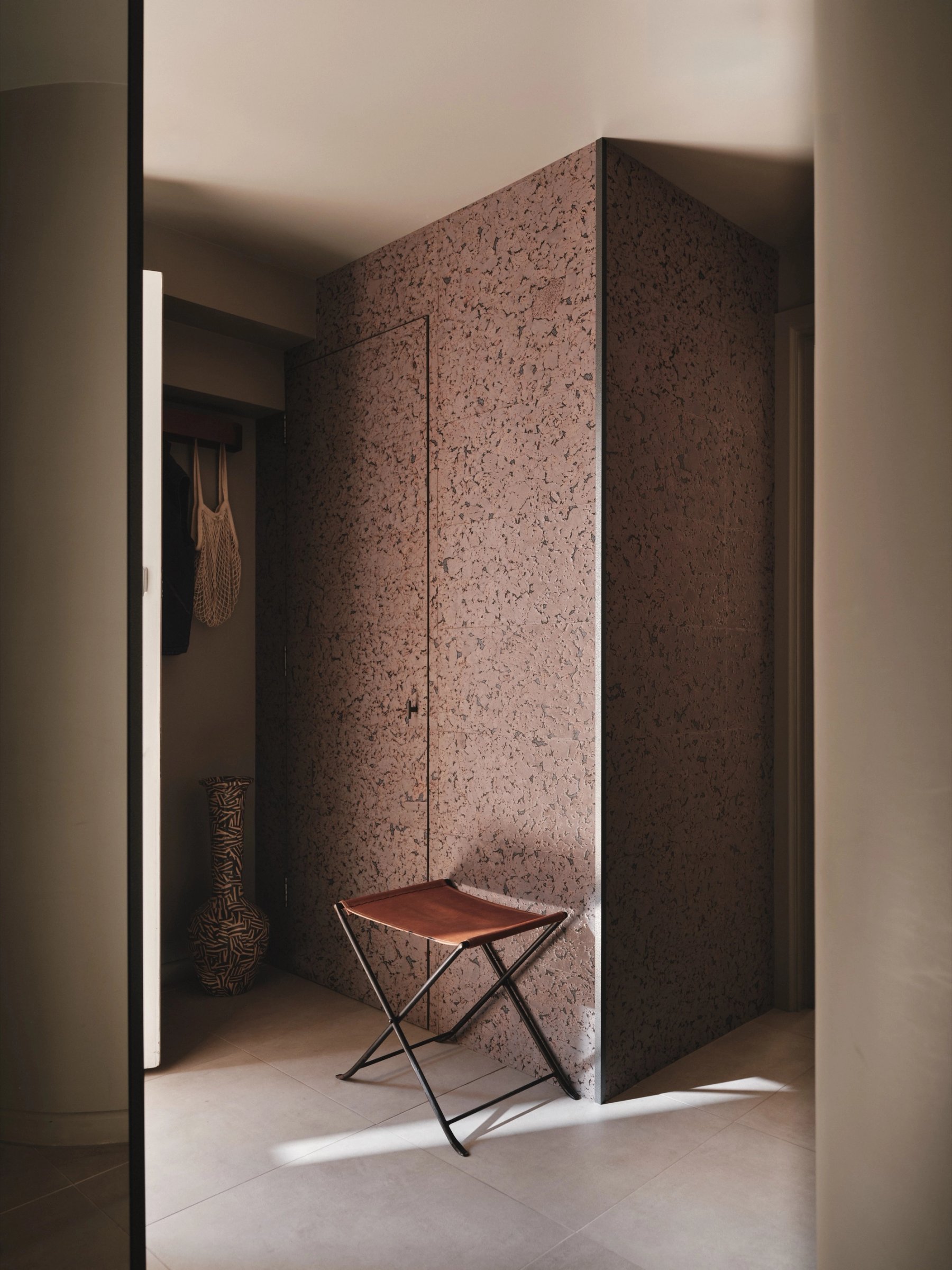 Hallway with a full-height storage volume clad in speckled pink-brown panelling, its door line almost invisible, a folding stool set against it.