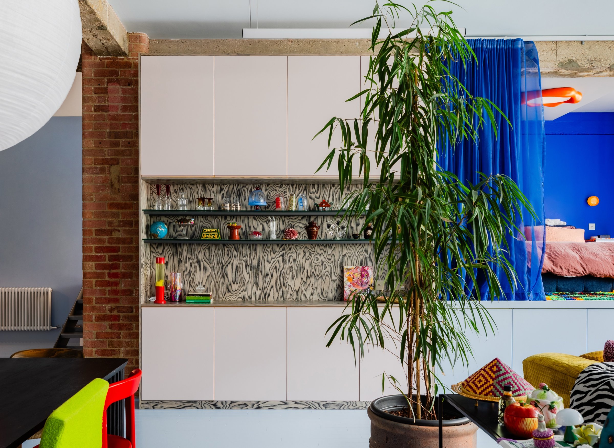 Warehouse living space with full-height pale pink cabinetry, its open shelf recess lined in figured ply, beside an exposed brick pier and concrete beam.