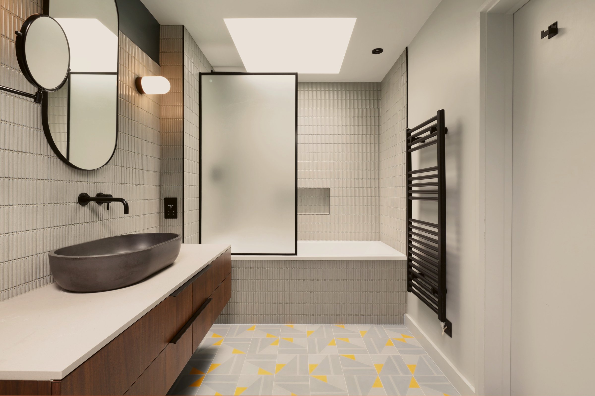 Bathroom from the door: walnut vanity and dark stone basin below a capsule mirror, bath and fluted glass screen beyond, geometric tiled floor throughout.