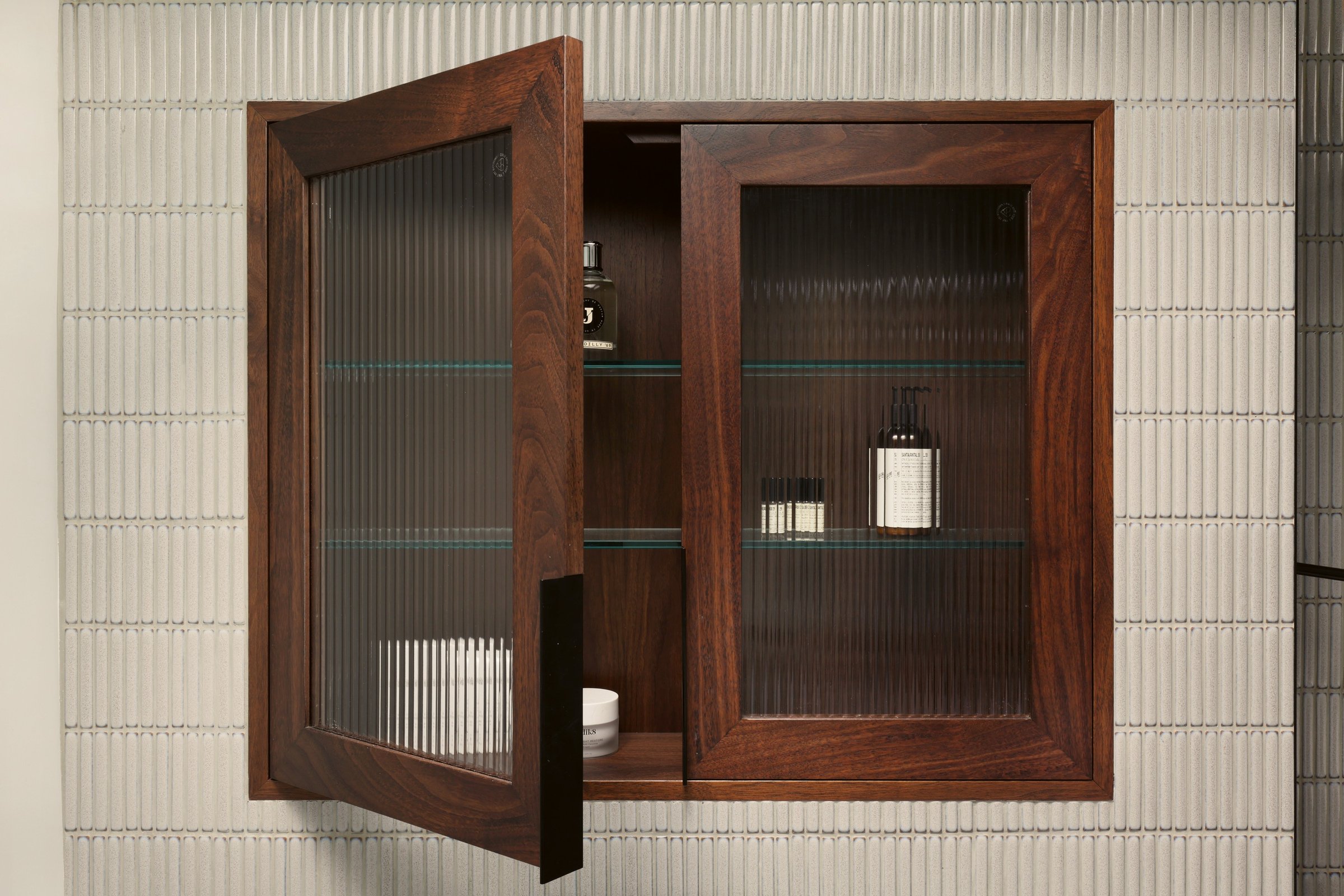 Walnut bathroom wall cabinet with one reeded glass door swung open on a slim black handle, glass shelves inside, against fluted white tiling.