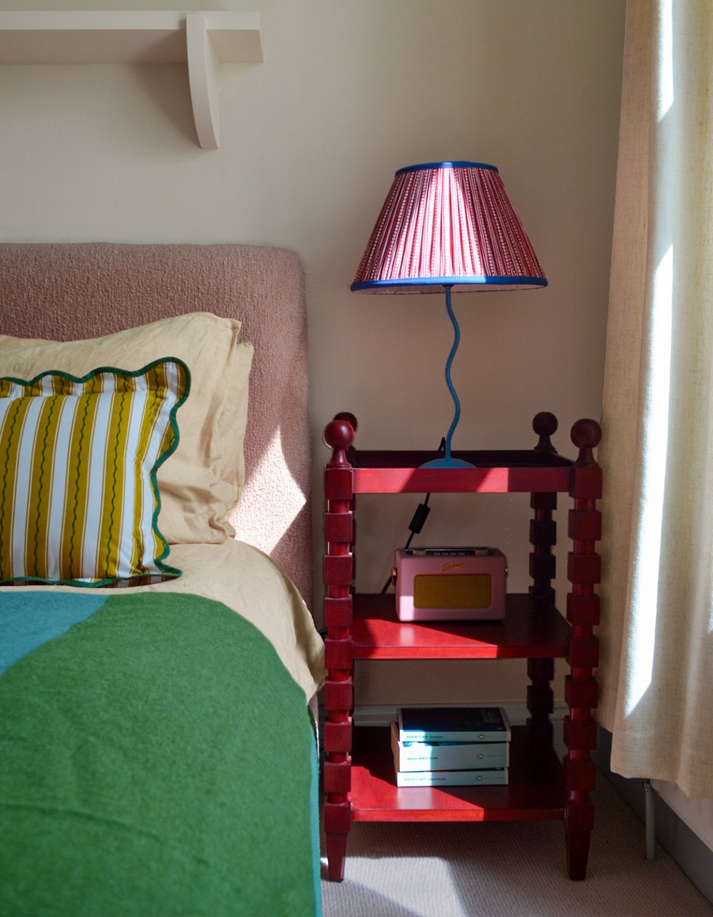 Bedroom detail with a red painted bobbin bedside stand, a pink pleated lampshade and striped cushions against a green blanket