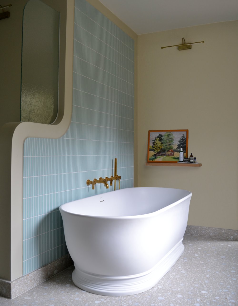 Freestanding roll-top bath against a panel of pale blue vertical tiles, with a brass floor-standing tap and a small framed landscape above