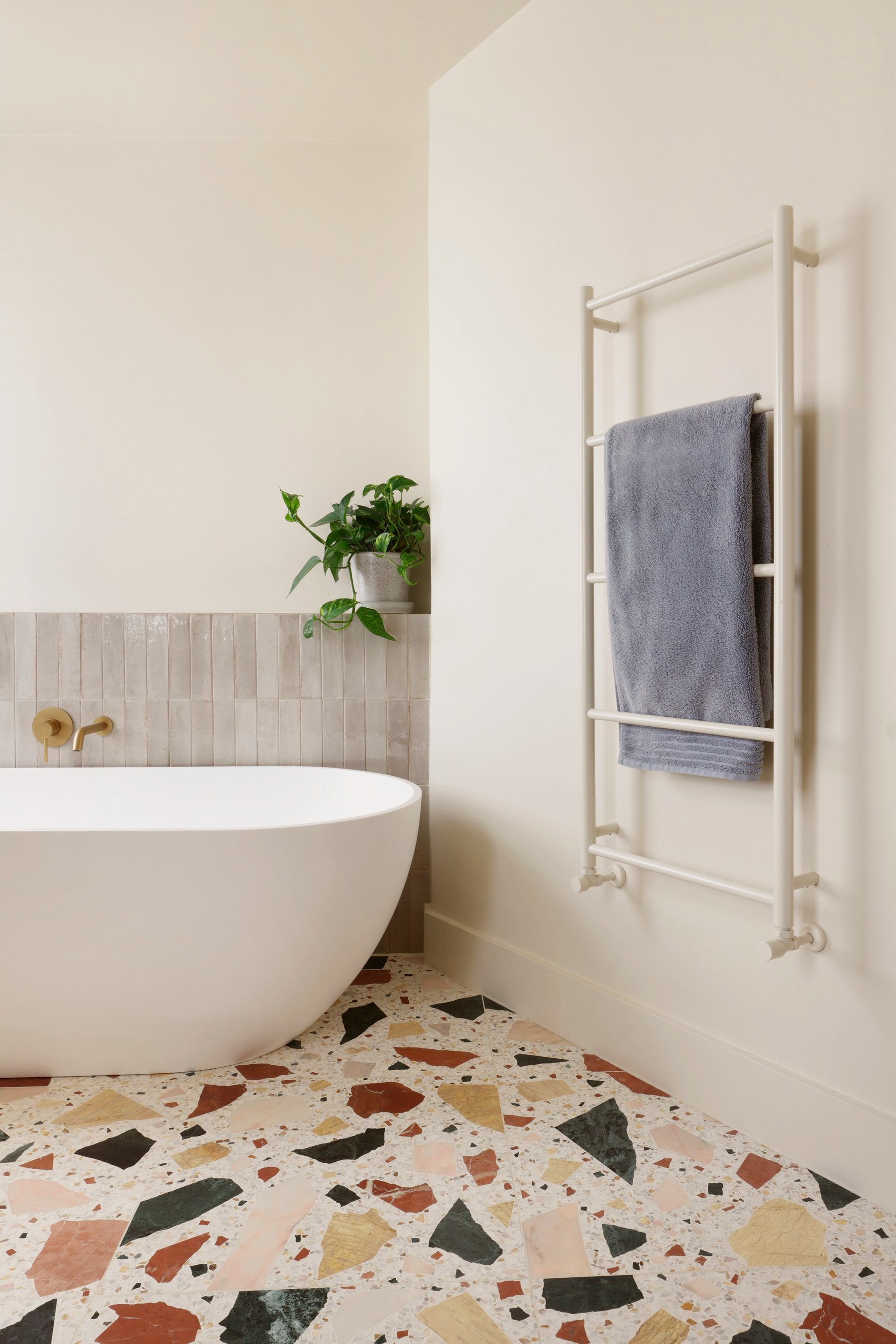 Bath end of the bathroom with a brass wall filler, a plant on the tiled shelf and a traditional towel rail holding a blue-grey towel.