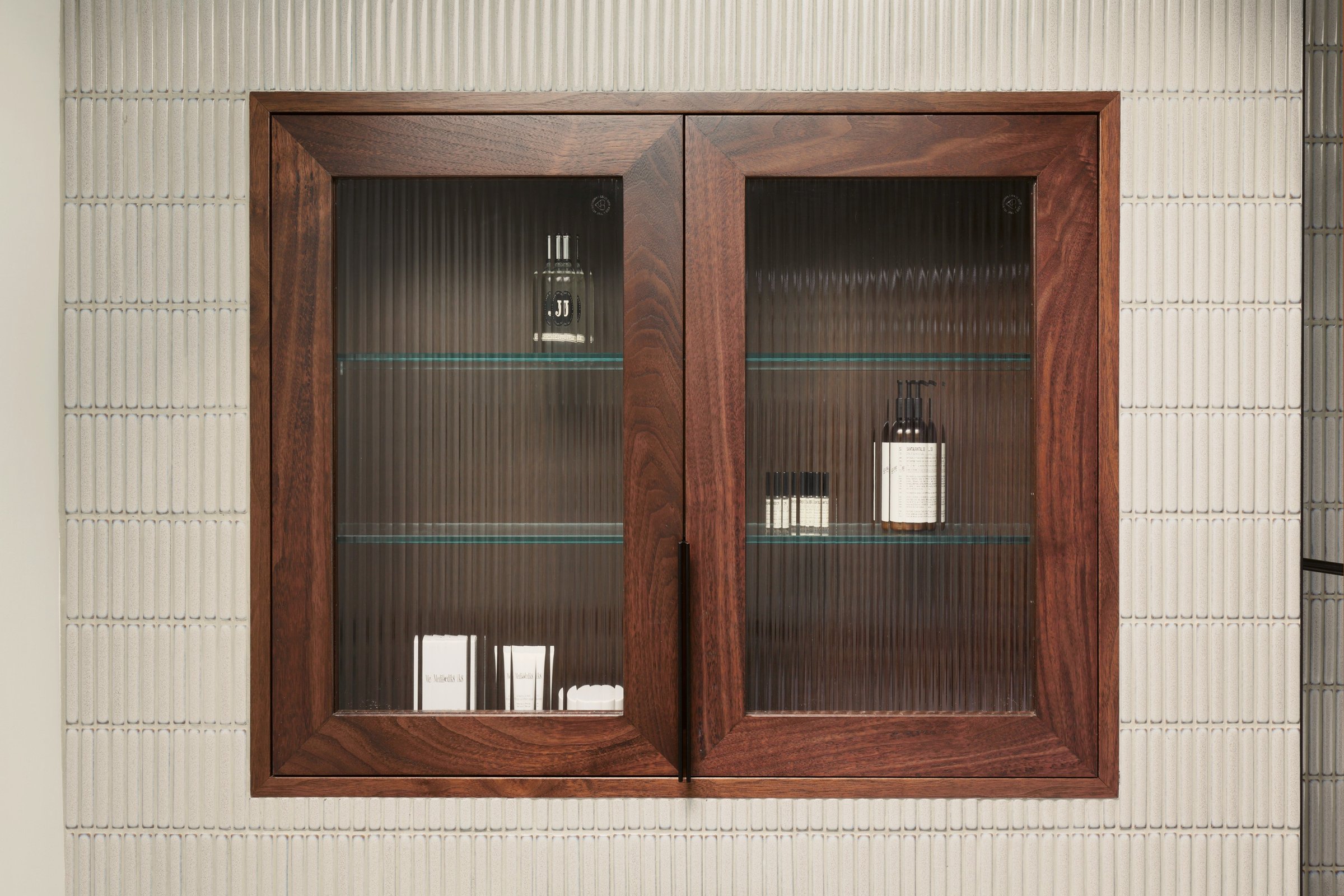 The walnut and reeded glass bathroom cabinet closed, its doors reducing the bottles behind to soft vertical bands, on fluted white tile.