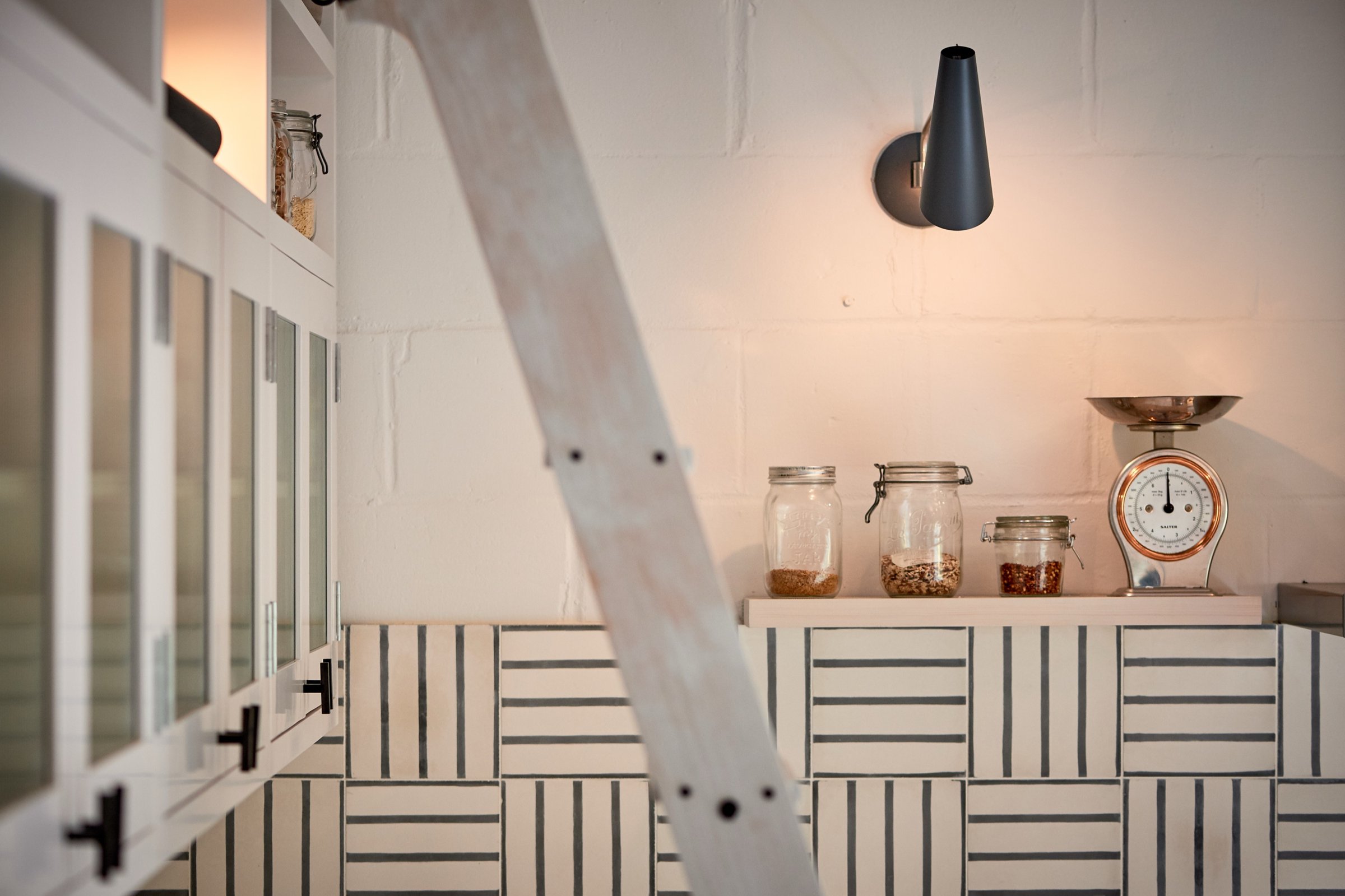 Kitchen corner with storage jars and vintage scales under a black cone wall light, and a limed timber library ladder leaning across the frame.