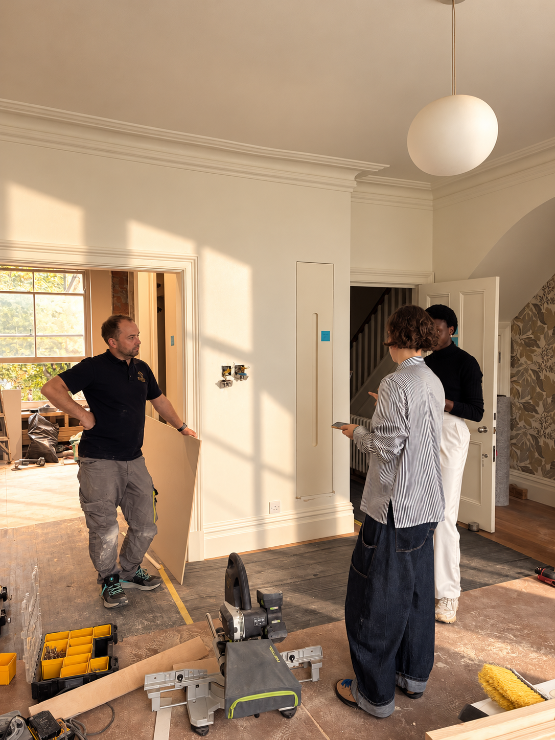 On site mid-renovation: a builder holding a sheet of board talks with two others in a sunlit period room, a mitre saw and tool case on the protected floor
