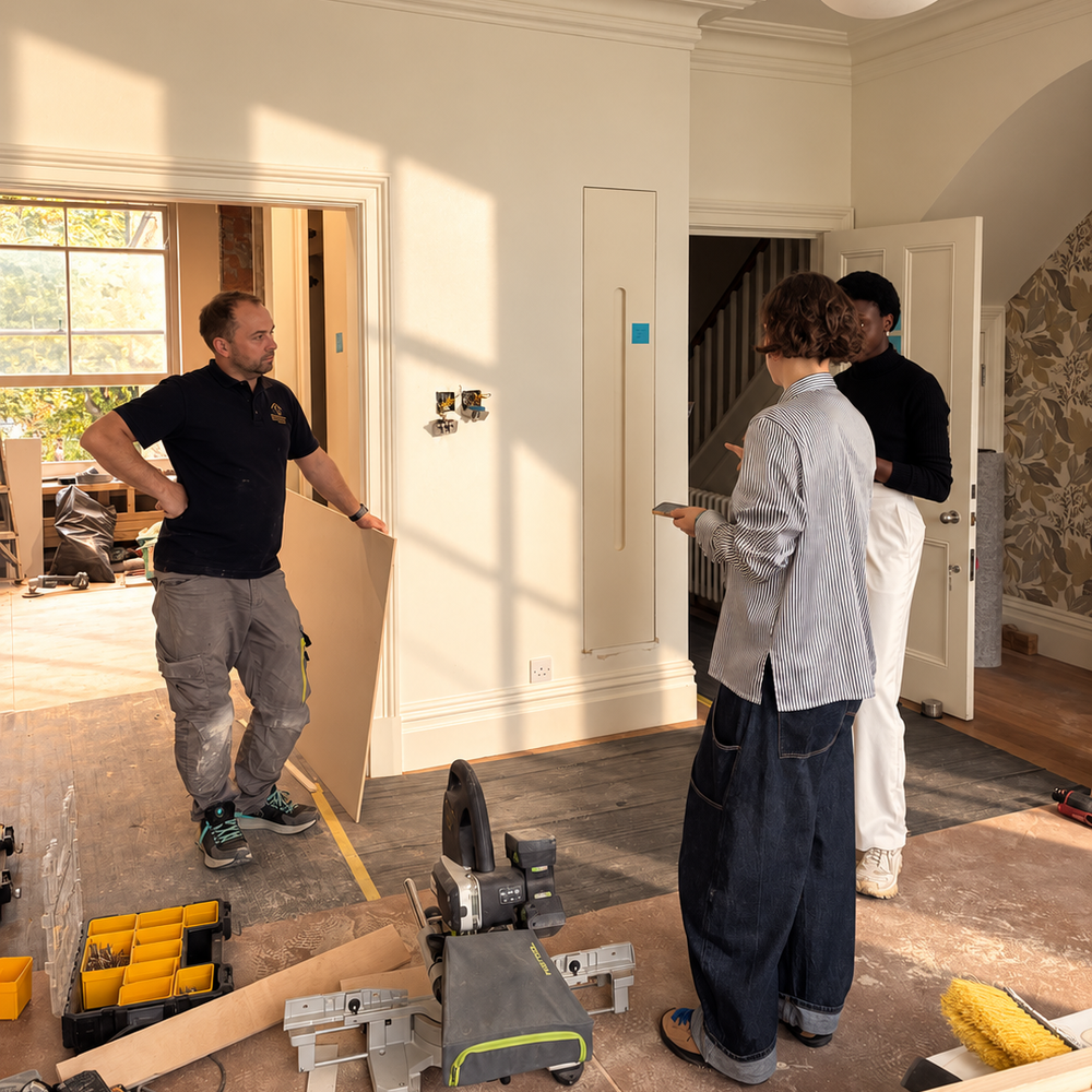 On site mid-renovation: a builder holding a sheet of board talks with two others in a sunlit period room, a mitre saw and tool case on the protected floor