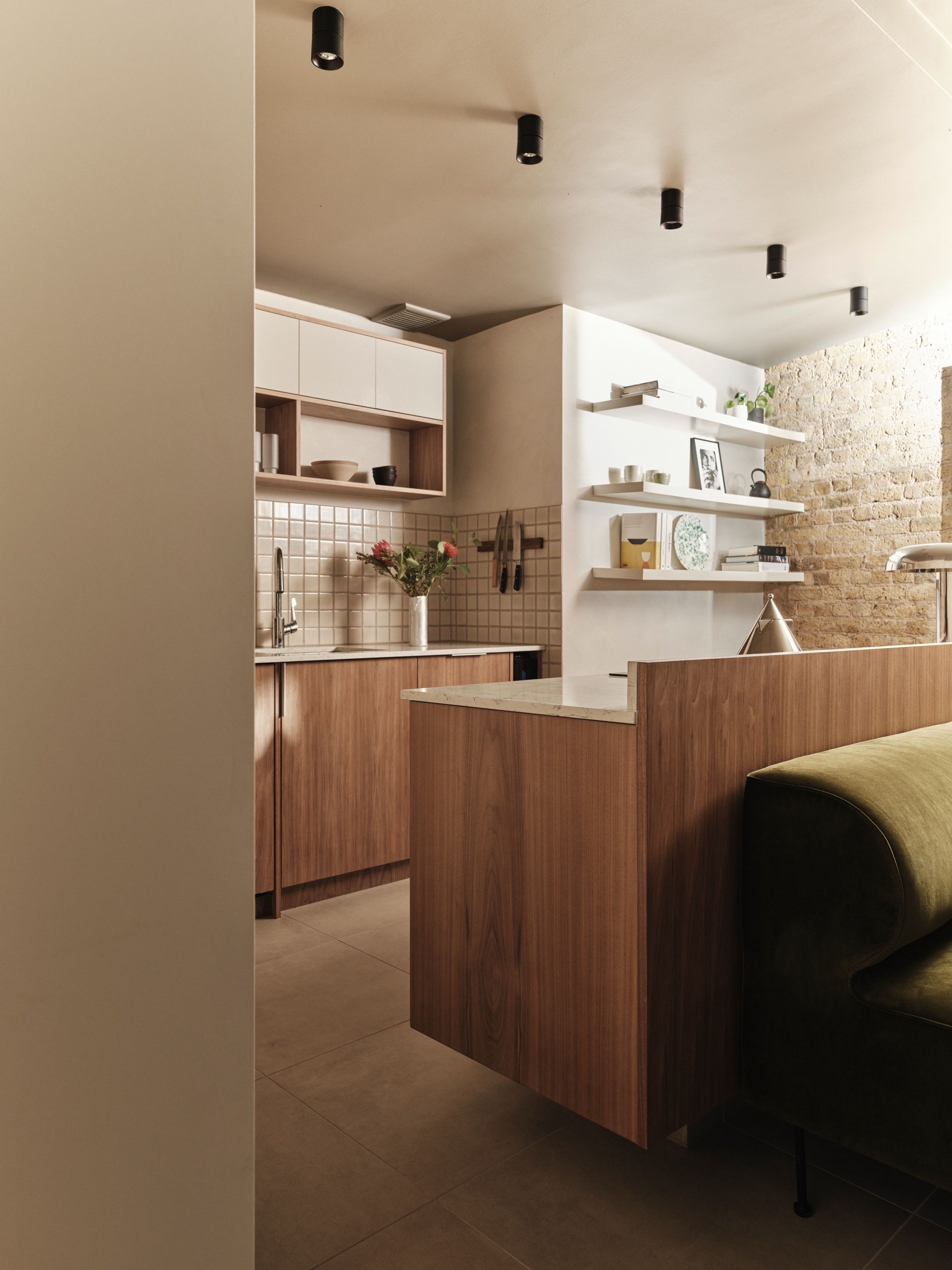 The walnut kitchen peninsula from the doorway, grain-matched and cantilevered clear of the floor, with the tiled sink run beyond.