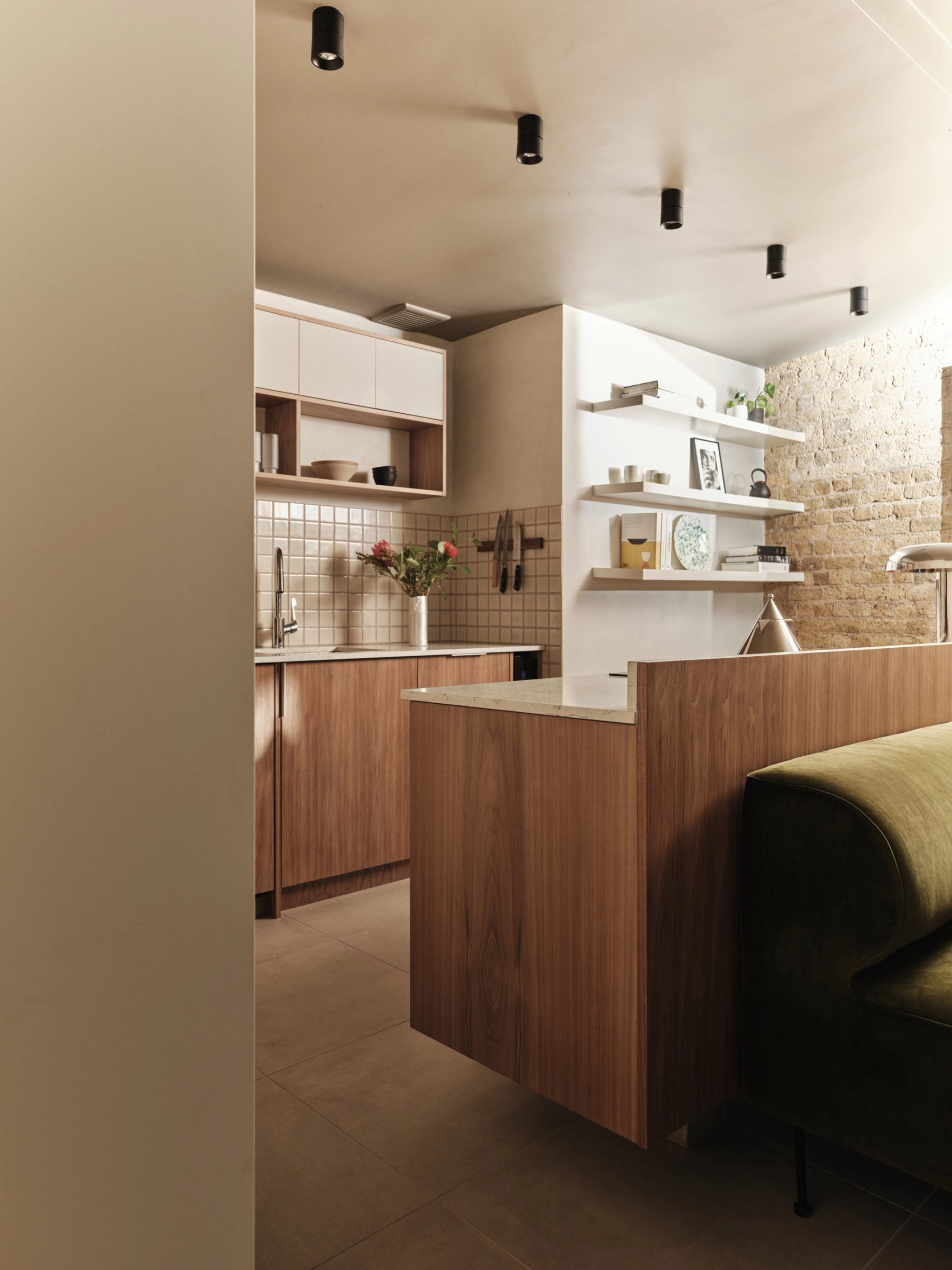 The walnut kitchen peninsula from the doorway, grain-matched and cantilevered clear of the floor, with the tiled sink run beyond.